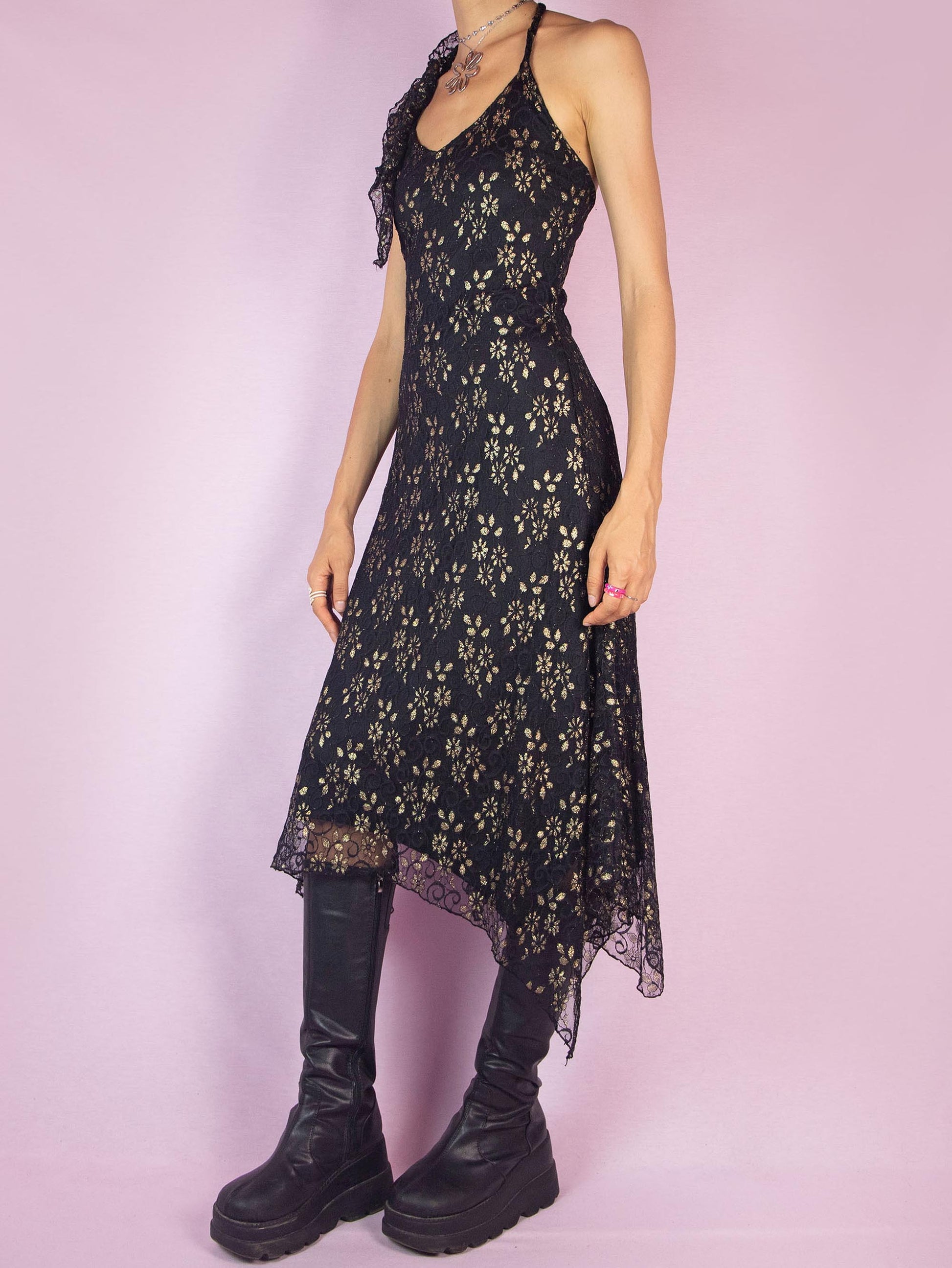 The Y2K Black Lace Asymmetrical Midi Dress is a vintage 2000s piece featuring a halter neck design and intricate golden floral lace detailing over a delicate mesh fabric. The dress is accented with ruffles along the neckline, adding a touch of romance and elegance. Perfect for a night out or special occasion.