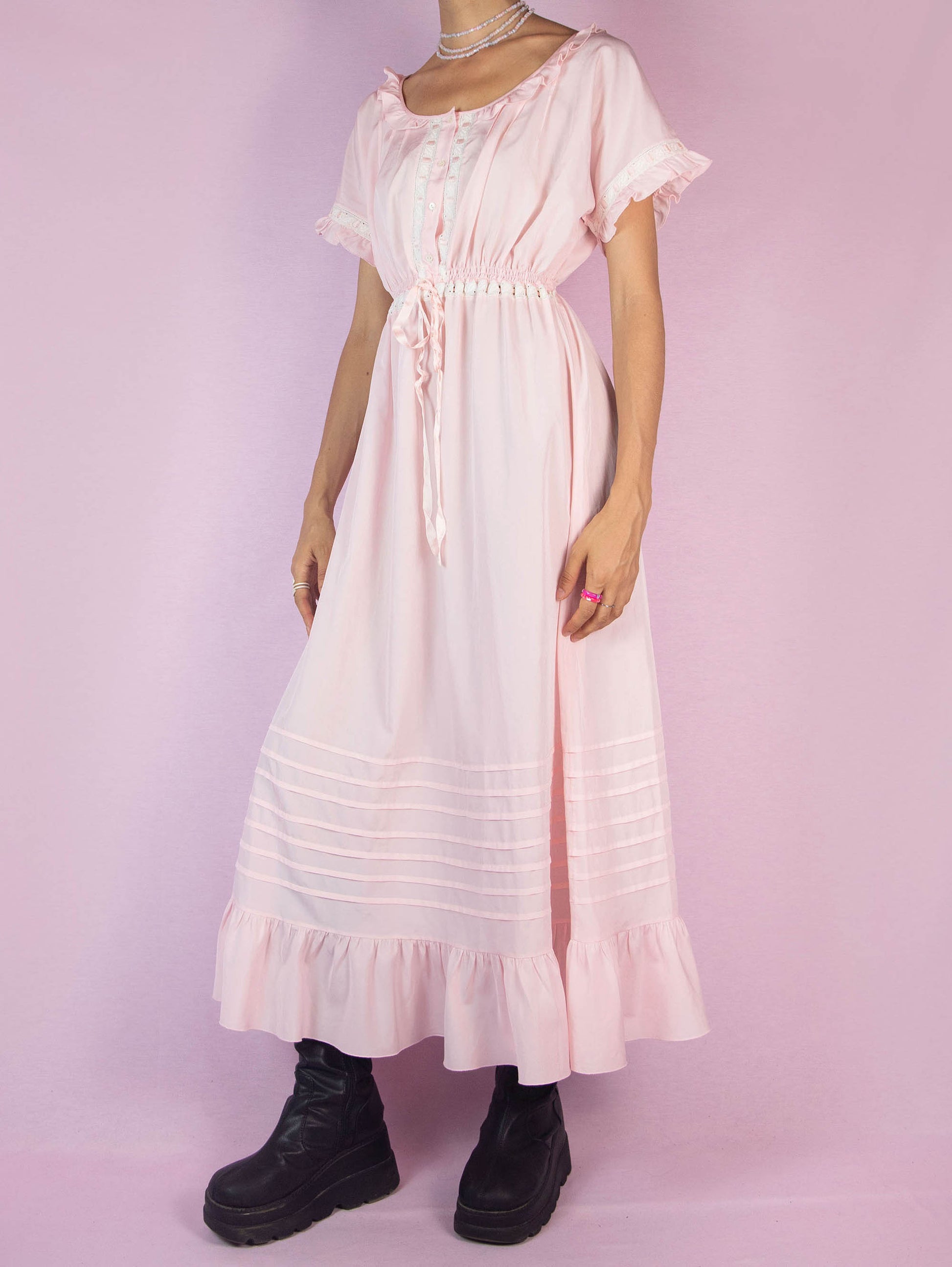 The Vintage 90s Light Pink Ruffle Midi Dress is a charming piece featuring a delicate ruffled neckline, short sleeves with lace trim, a buttoned front, a cinched waist with a ribbon tie, and a flowing midi-length skirt with a ruffle hem. Perfect for casual outings or lounging at home.