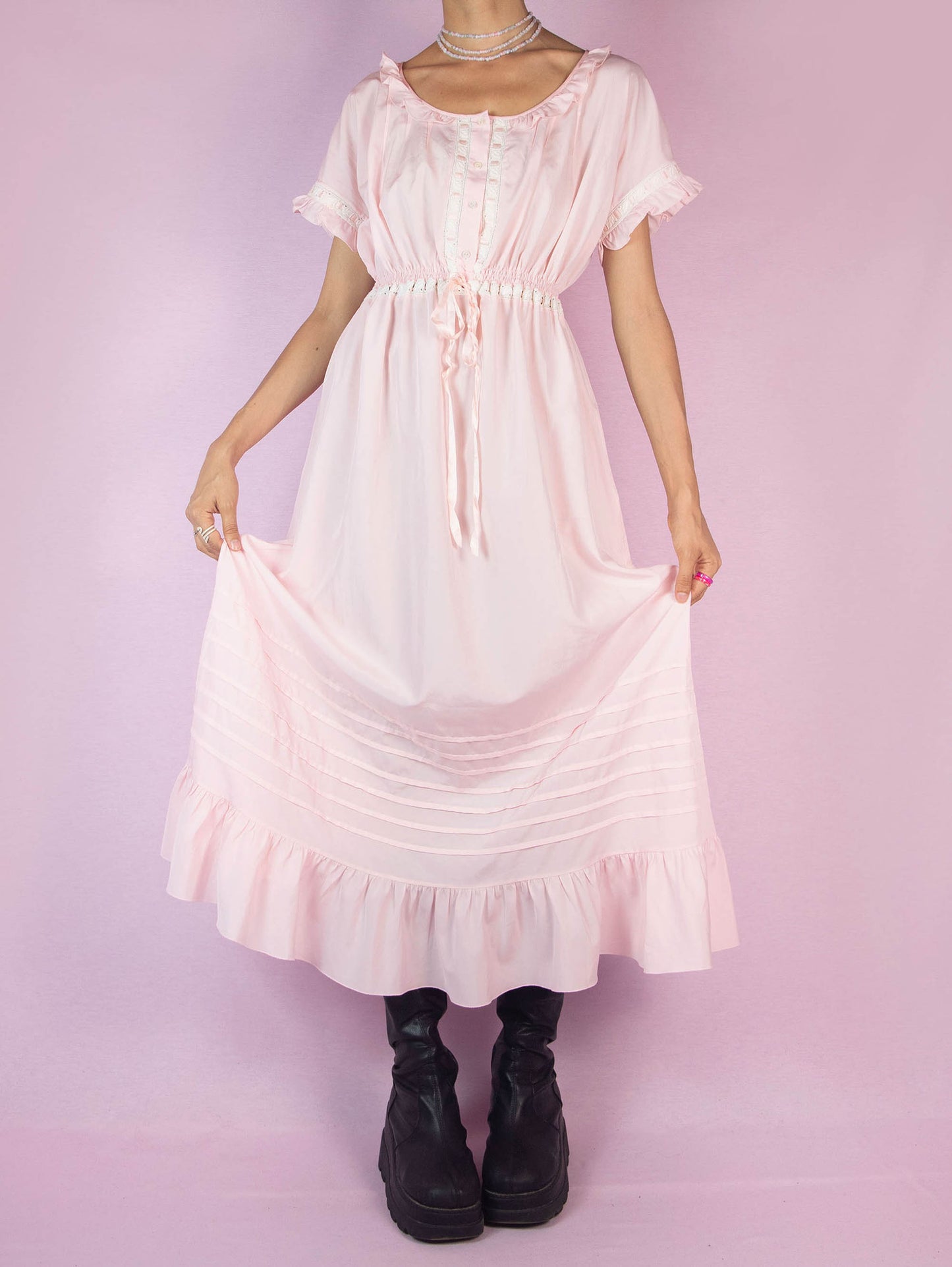 The Vintage 90s Light Pink Ruffle Midi Dress is a charming piece featuring a delicate ruffled neckline, short sleeves with lace trim, a buttoned front, a cinched waist with a ribbon tie, and a flowing midi-length skirt with a ruffle hem. Perfect for casual outings or lounging at home.