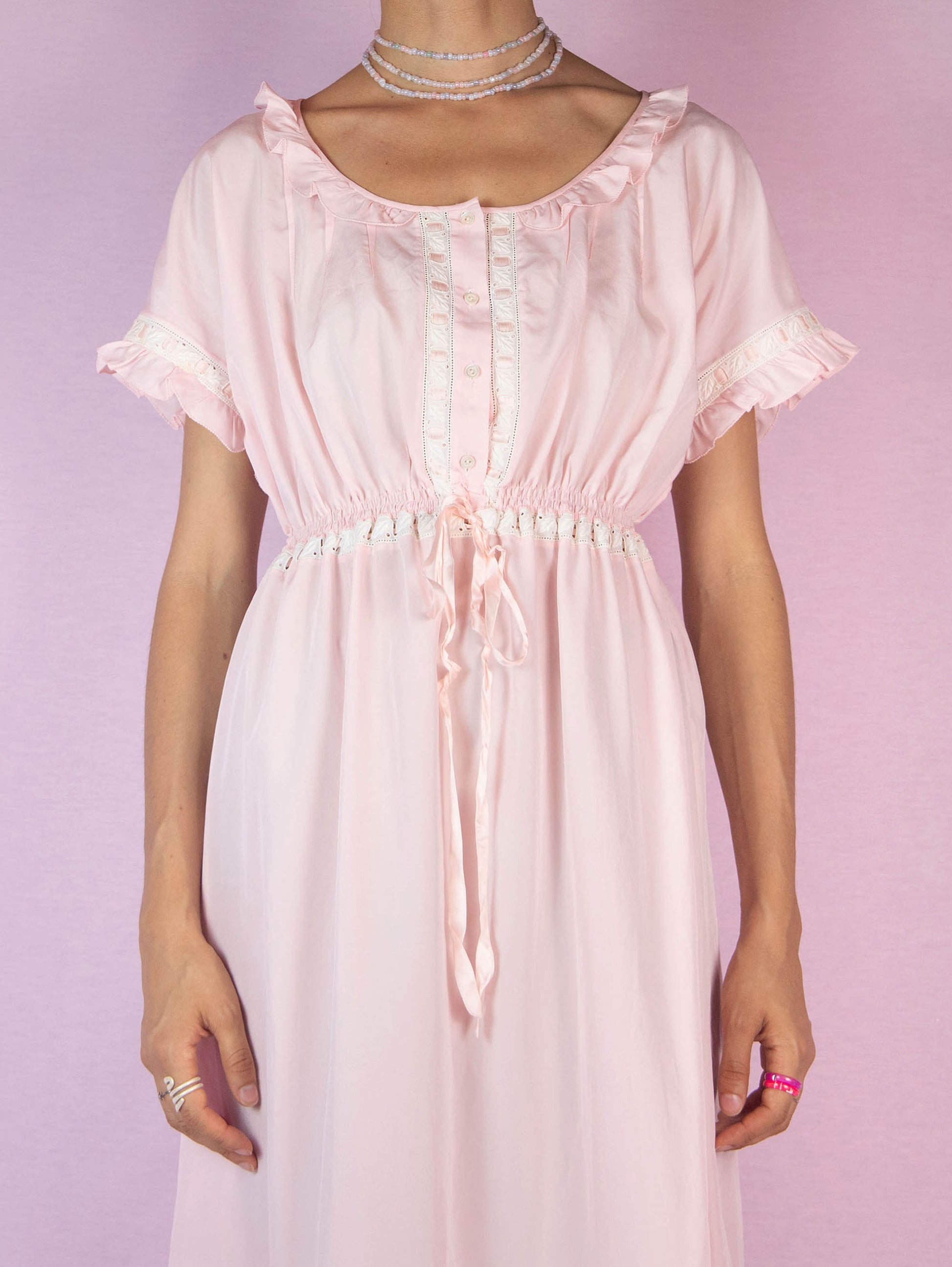 The Vintage 90s Light Pink Ruffle Midi Dress is a charming piece featuring a delicate ruffled neckline, short sleeves with lace trim, a buttoned front, a cinched waist with a ribbon tie, and a flowing midi-length skirt with a ruffle hem. Perfect for casual outings or lounging at home.