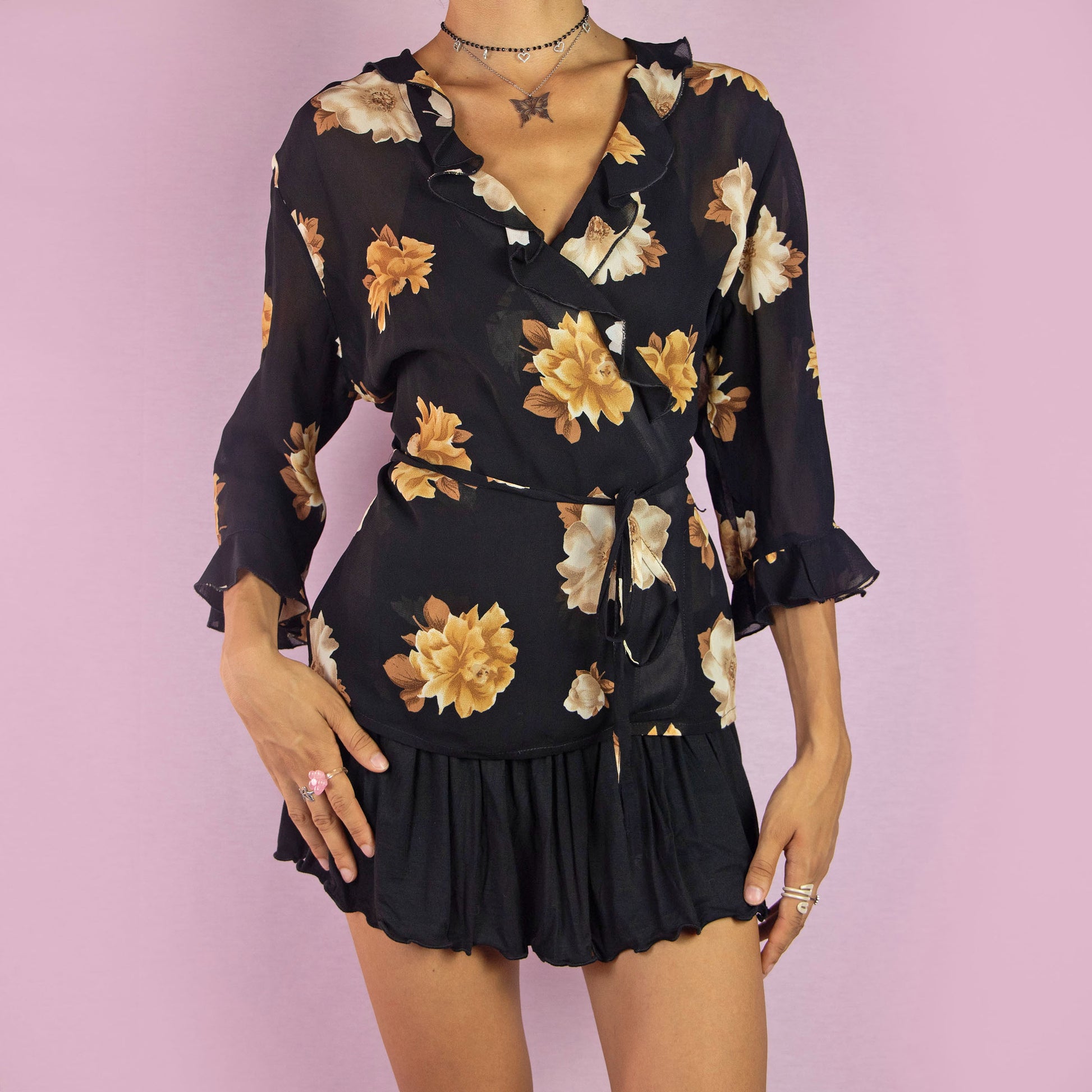 The Vintage 90s Black Ruffle Tie Wrap Top is a blouse featuring a semi-sheer floral print, three-quarter sleeves, a ruffled neckline, and a wrap tie waist. Perfect for a casual day out or a more dressed-up occasion.