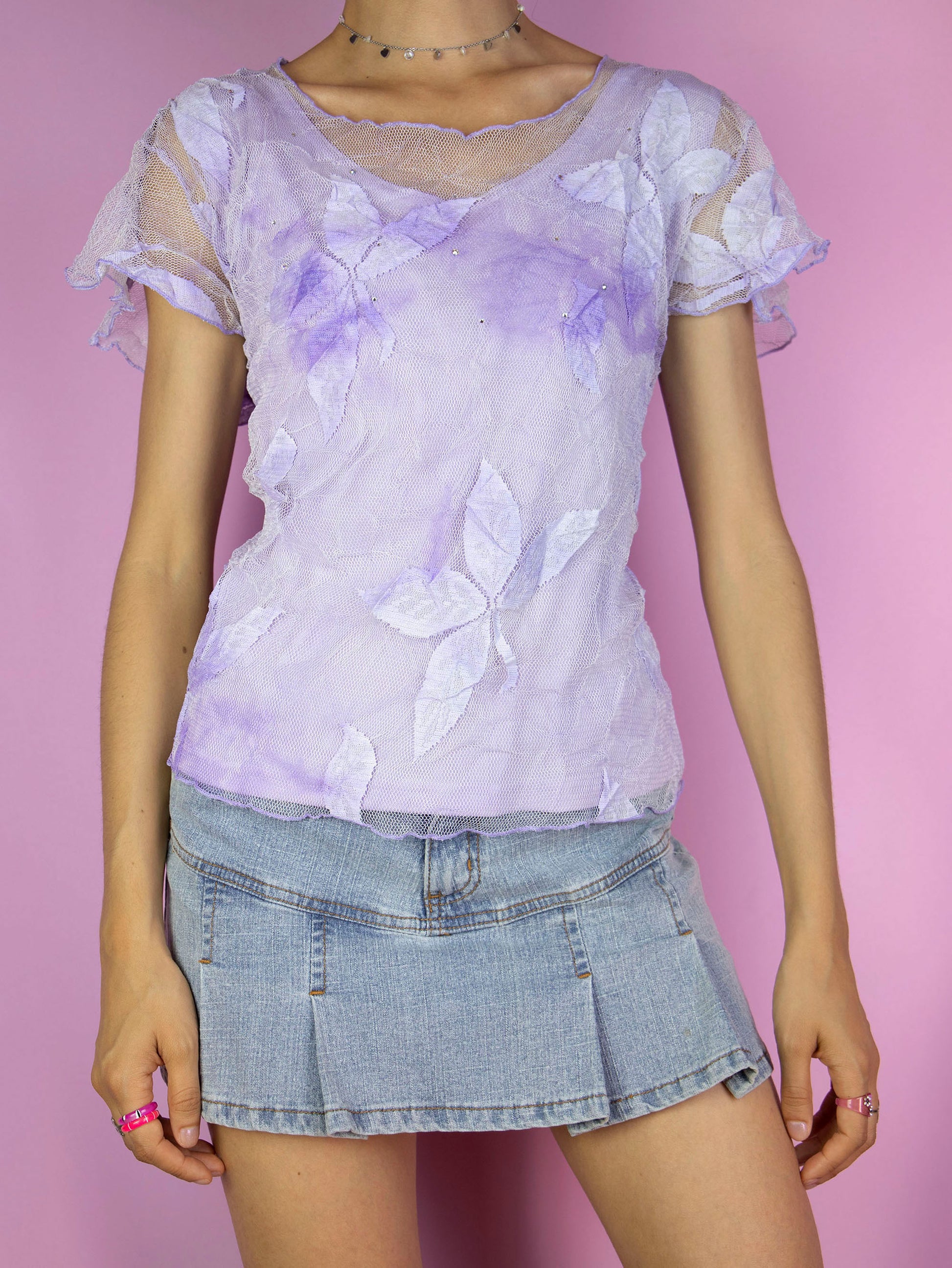 The Y2K Lilac Mesh Sheer Top is a vintage short-sleeve, pastel purple semi-sheer mesh top adorned with delicate floral tulle and rhinestones. Romantic coquette 2000s fairy grunge shirt.