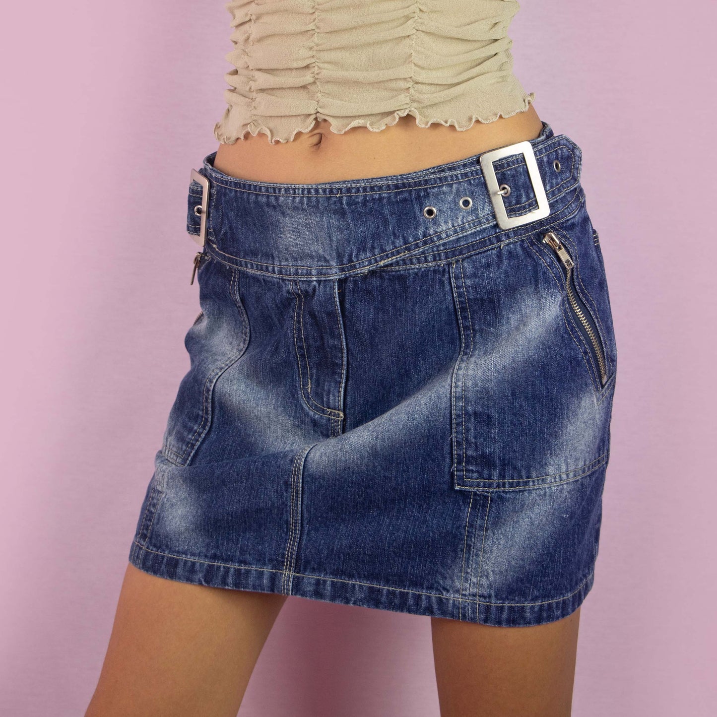 The Y2K Denim Buckle Mini Skirt is a vintage 2000s piece featuring a mid-rise fit and a structured A-line silhouette. Made from medium-wash denim, this skirt has unique buckle details at the waist, exposed zipper accents, front pockets and a back zipper closure. Perfect for casual wear and parties.