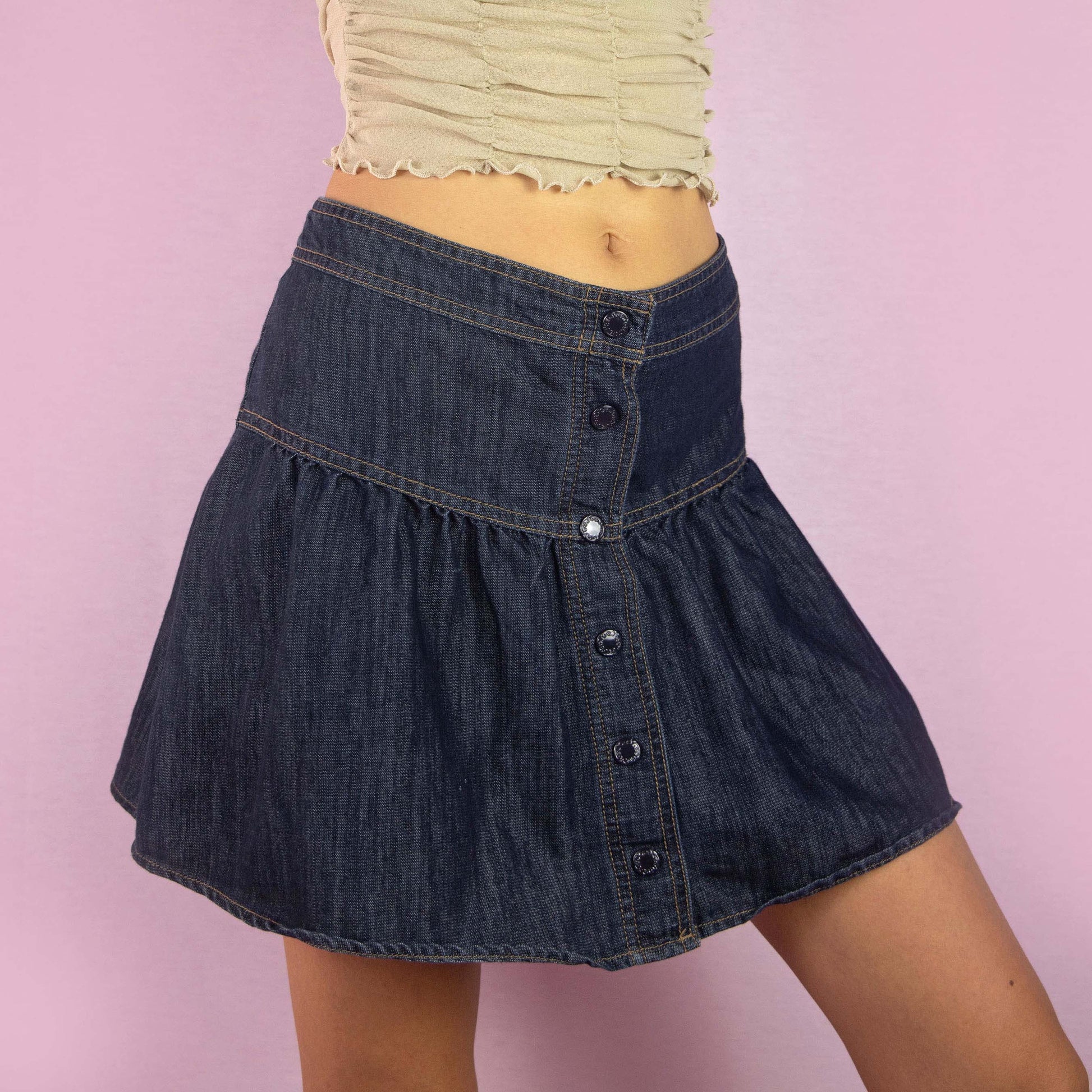 The Y2K Denim Flare Mini Skirt is a vintage 2000s piece featuring a mid-rise fit and a flared A-line silhouette. Made from dark-wash denim, this skirt showcases a tiered design with button-up detailing along the front. Ideal for casual wear and summer outings.