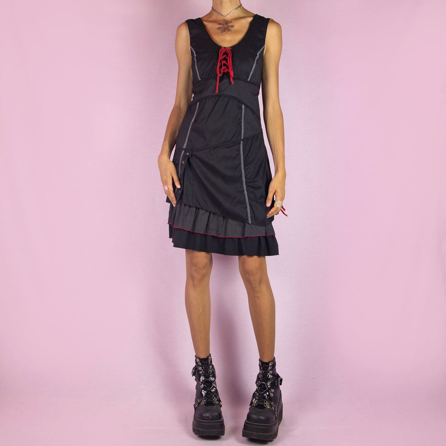 The Y2K Black Layered Lace-Up Dress is a vintage 2000s mini dress featuring a sleeveless design, scoop neckline, and stretchy fabric. It showcases a red lace-up detail at the bodice, contrasting stitching throughout, and a side zipper closure. The layered skirt, with its pinstripe and solid panels, button accents, and ruffle hem, adds a touch of whimsy. Perfect for night events and parties.