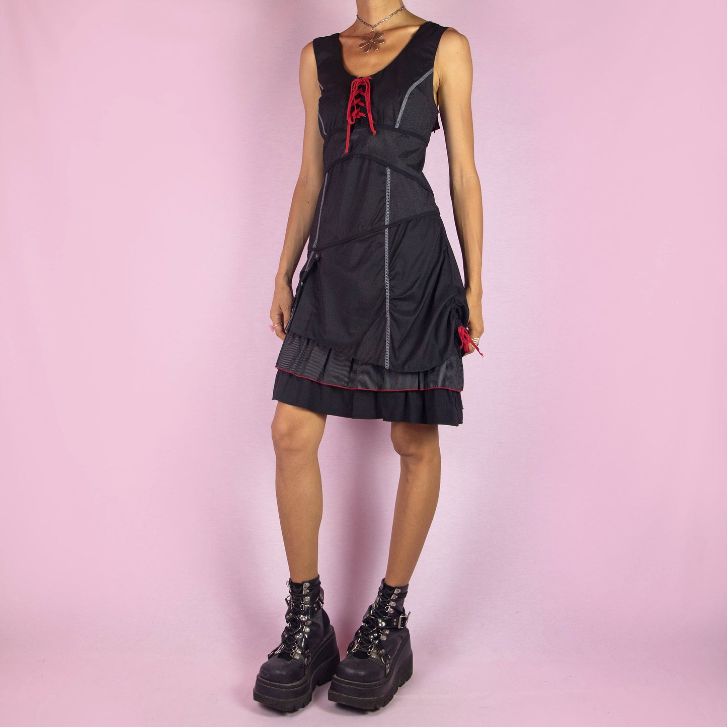 The Y2K Black Layered Lace-Up Dress is a vintage 2000s mini dress featuring a sleeveless design, scoop neckline, and stretchy fabric. It showcases a red lace-up detail at the bodice, contrasting stitching throughout, and a side zipper closure. The layered skirt, with its pinstripe and solid panels, button accents, and ruffle hem, adds a touch of whimsy. Perfect for night events and parties.