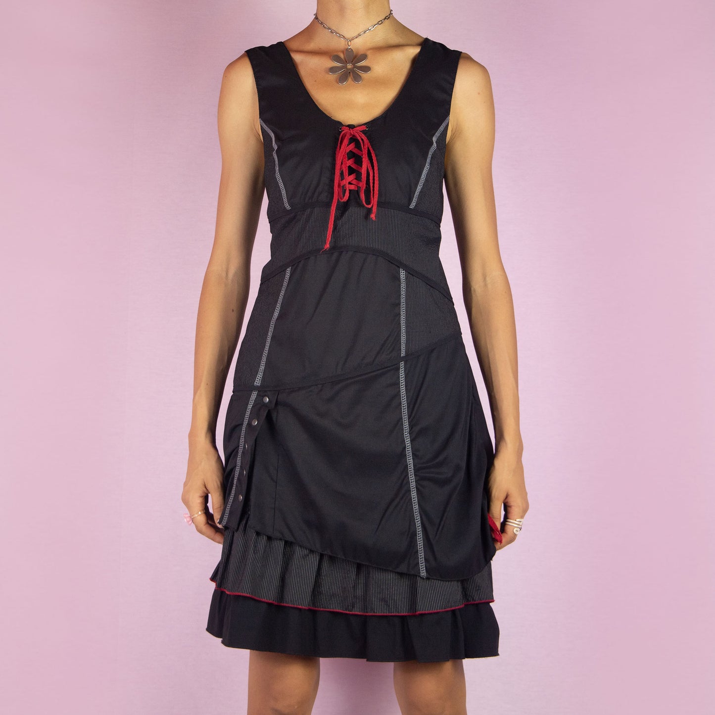 The Y2K Black Layered Lace-Up Dress is a vintage 2000s mini dress featuring a sleeveless design, scoop neckline, and stretchy fabric. It showcases a red lace-up detail at the bodice, contrasting stitching throughout, and a side zipper closure. The layered skirt, with its pinstripe and solid panels, button accents, and ruffle hem, adds a touch of whimsy. Perfect for night events and parties.