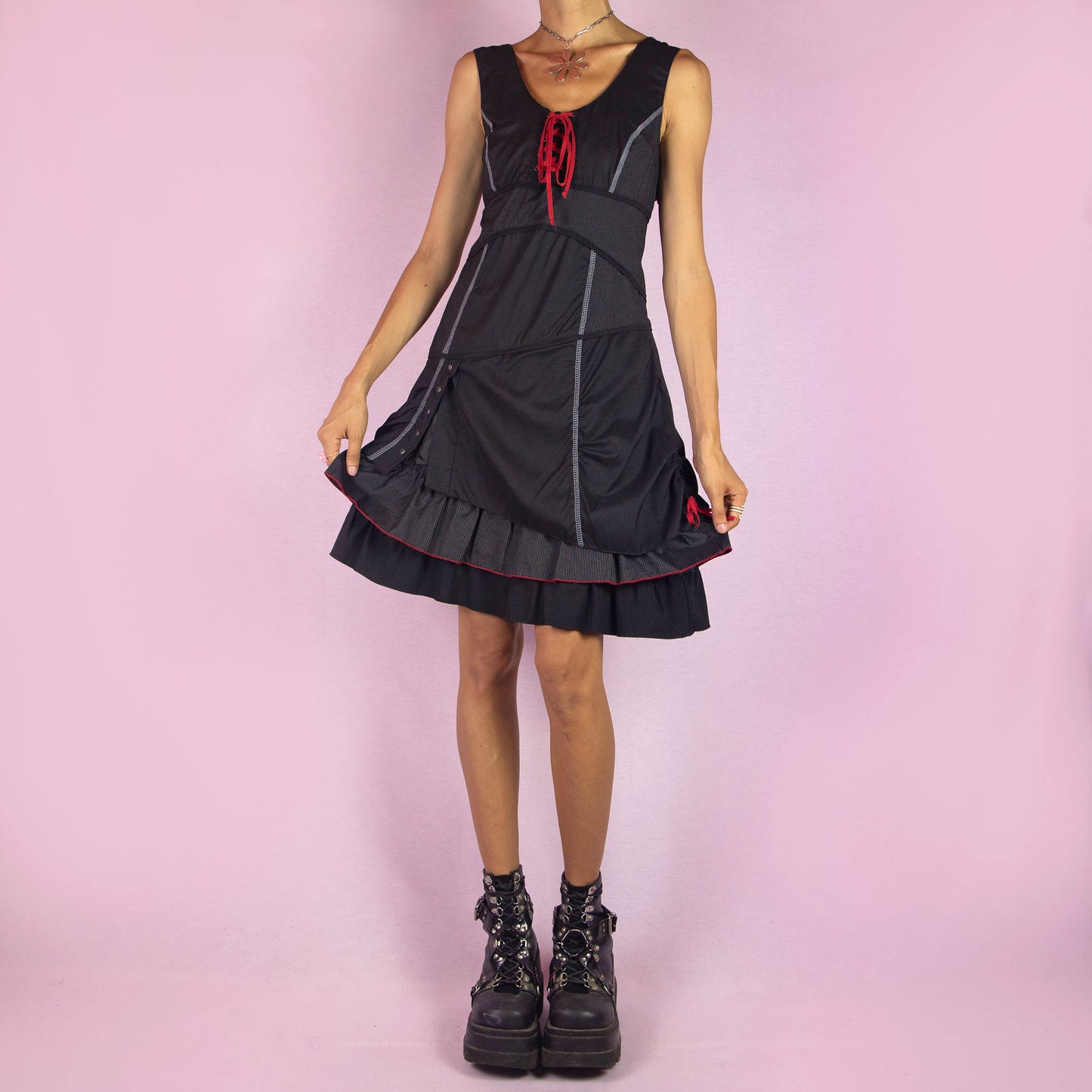 The Y2K Black Layered Lace-Up Dress is a vintage 2000s mini dress featuring a sleeveless design, scoop neckline, and stretchy fabric. It showcases a red lace-up detail at the bodice, contrasting stitching throughout, and a side zipper closure. The layered skirt, with its pinstripe and solid panels, button accents, and ruffle hem, adds a touch of whimsy. Perfect for night events and parties.