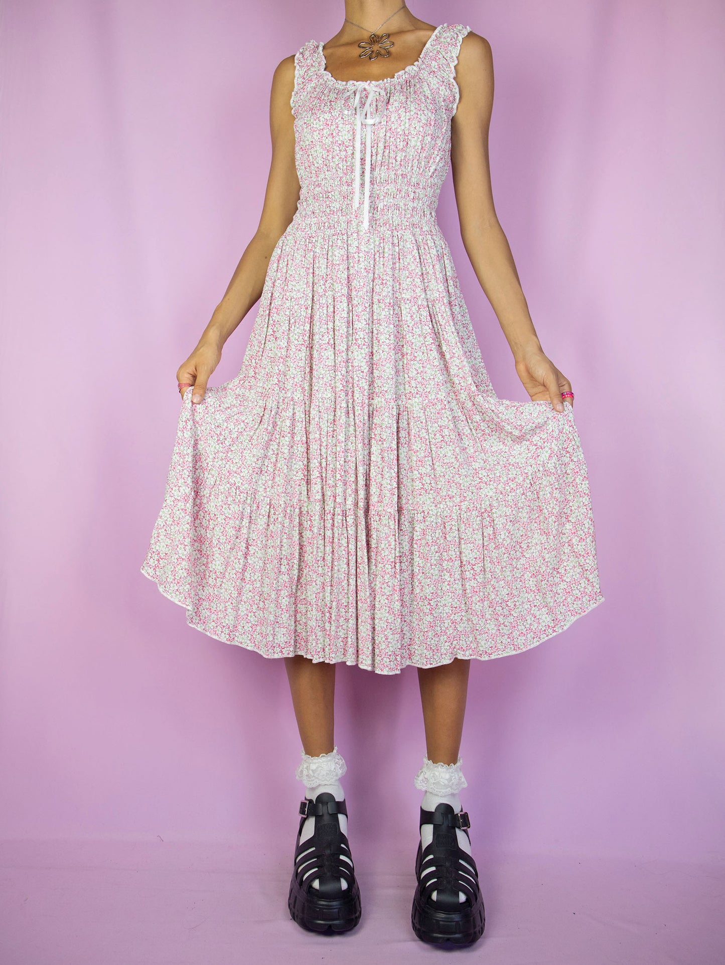 The Y2K Floral Milkmaid Midi Dress is a vintage multicolored floral pink and white sleeveless dress with tiered layers, ruched waist, and a bow at the front. This super cute cottage prairie-inspired boho summer dress is from the 2000s.