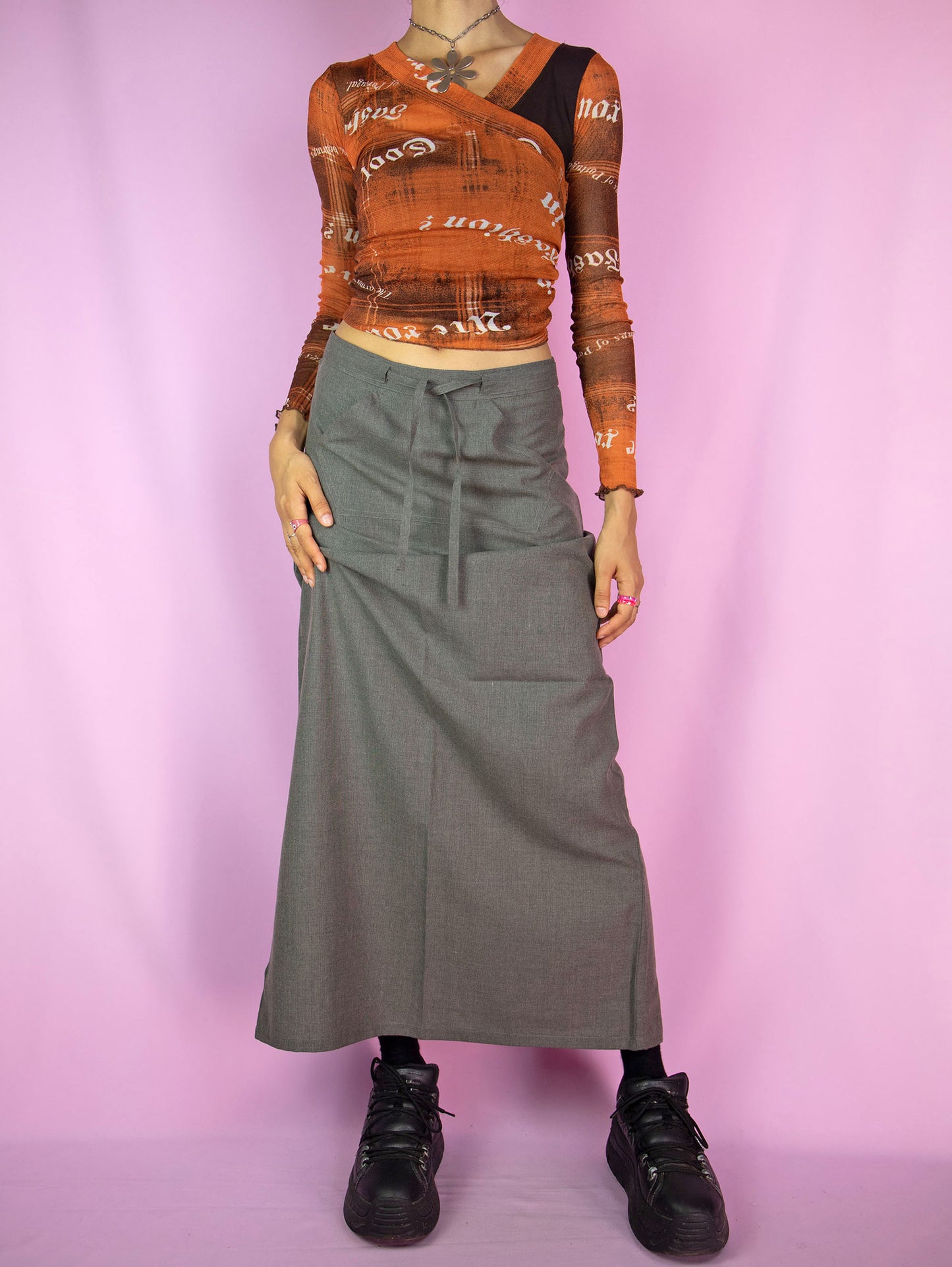 The Vintage 90s Gray Maxi Skirt features a mid-rise, A-line design with a tie-front detail, a front pocket, a back zipper closure, and a back slit. This longline skirt has a sleek, minimalist look.