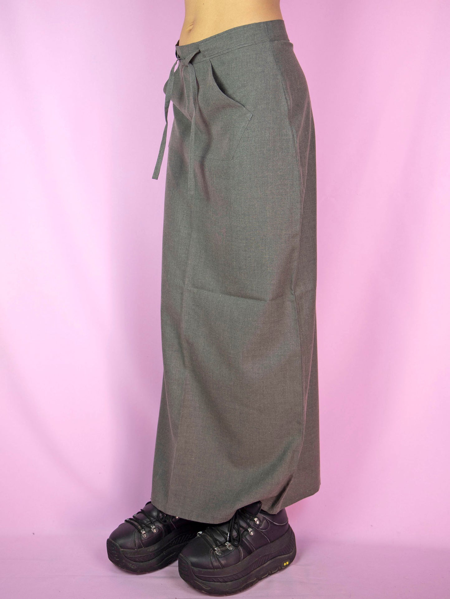 The Vintage 90s Gray Maxi Skirt features a mid-rise, A-line design with a tie-front detail, a front pocket, a back zipper closure, and a back slit. This longline skirt has a sleek, minimalist look.