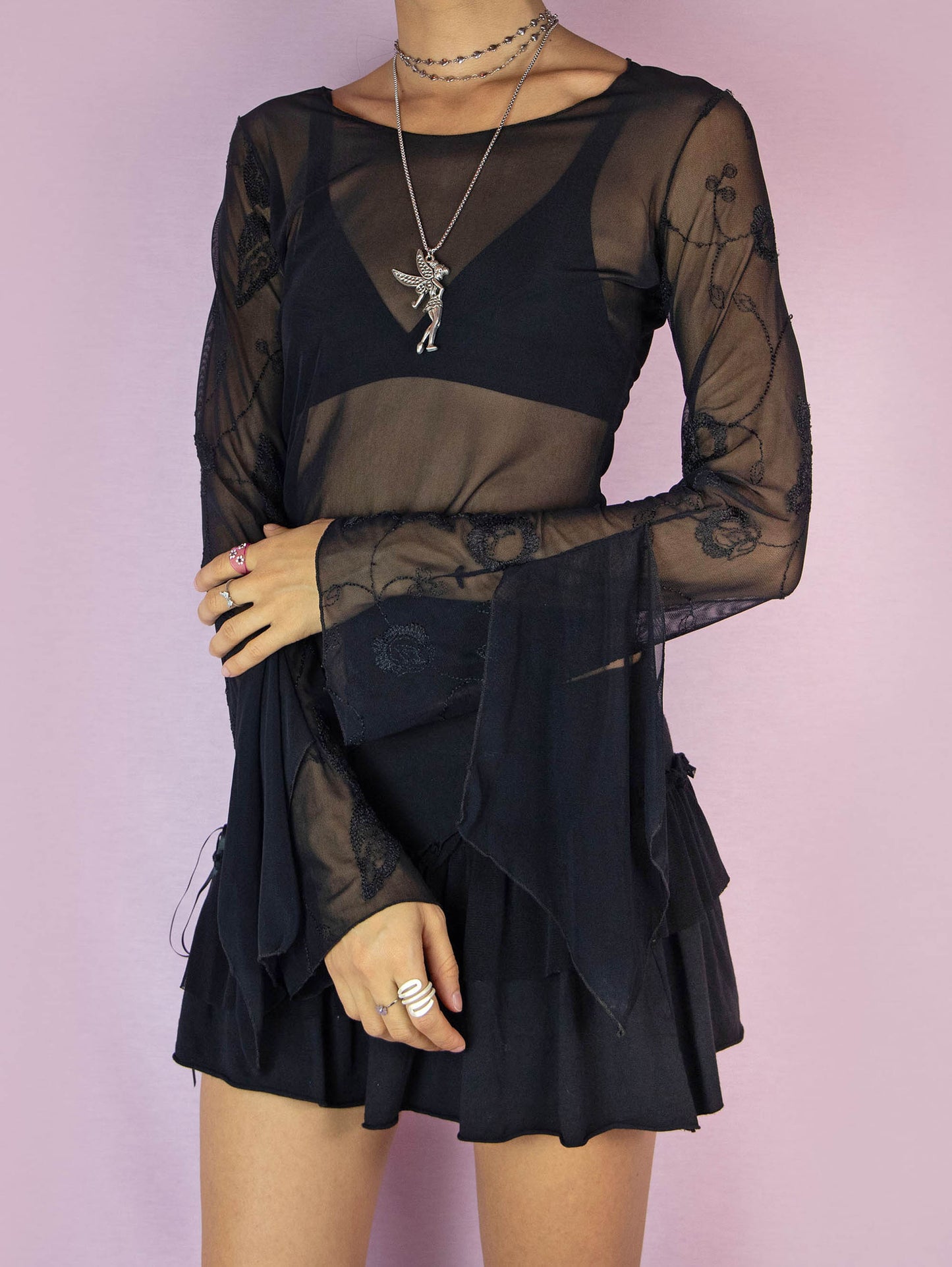 The Y2K Black Mesh Asymmetric Top is a vintage 2000s long sleeve semi-sheer night rave party shirt with bell sleeves, a pointed asymmetrical hem, and embroidered details.