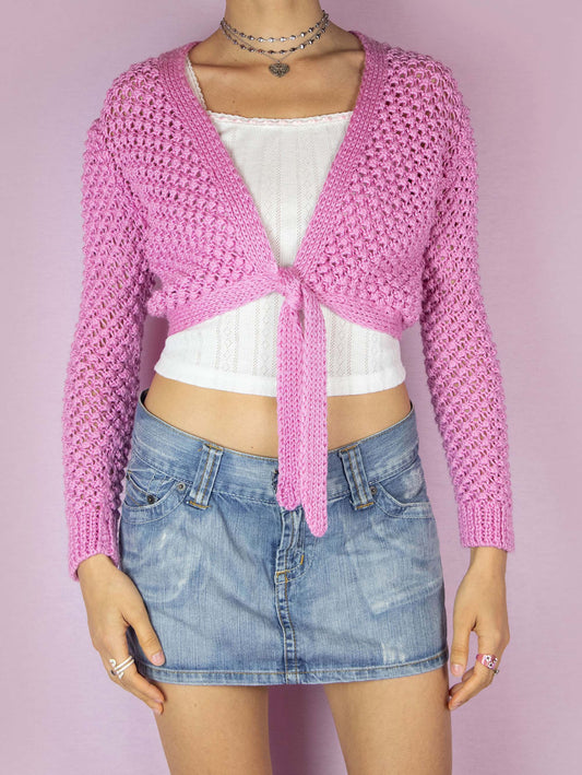 The Y2K Pink Knit Tie Cardigan is a vintage 2000s boho coquette inspired bolero jacket that features an open-knit design with long sleeves and a cropped length. It has a V-neckline and ties at the front for an adjustable fit. The vibrant pink color and textured knit make it a playful layering piece.