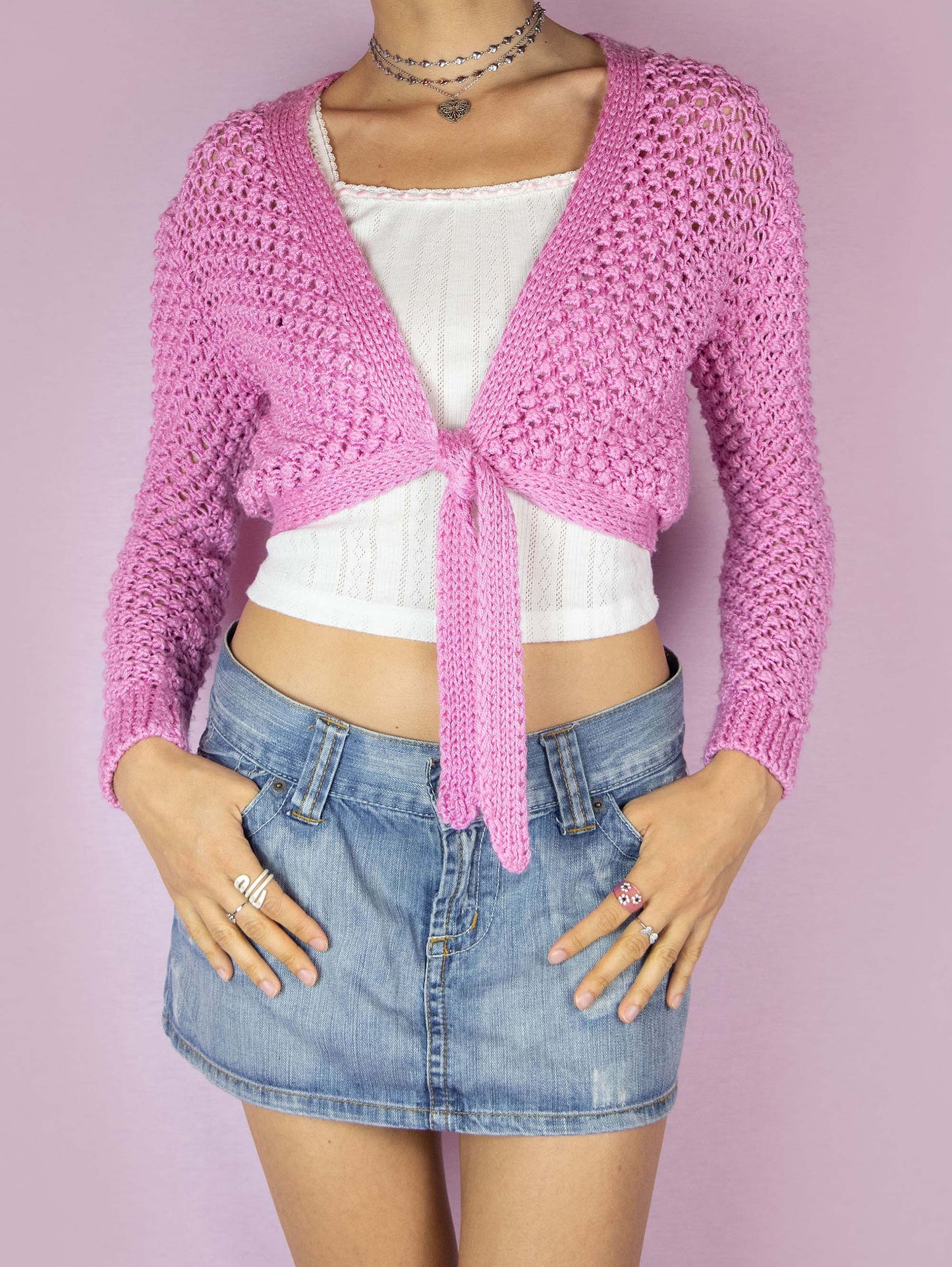 The Y2K Pink Knit Tie Cardigan is a vintage 2000s boho coquette inspired bolero jacket that features an open-knit design with long sleeves and a cropped length. It has a V-neckline and ties at the front for an adjustable fit. The vibrant pink color and textured knit make it a playful layering piece.