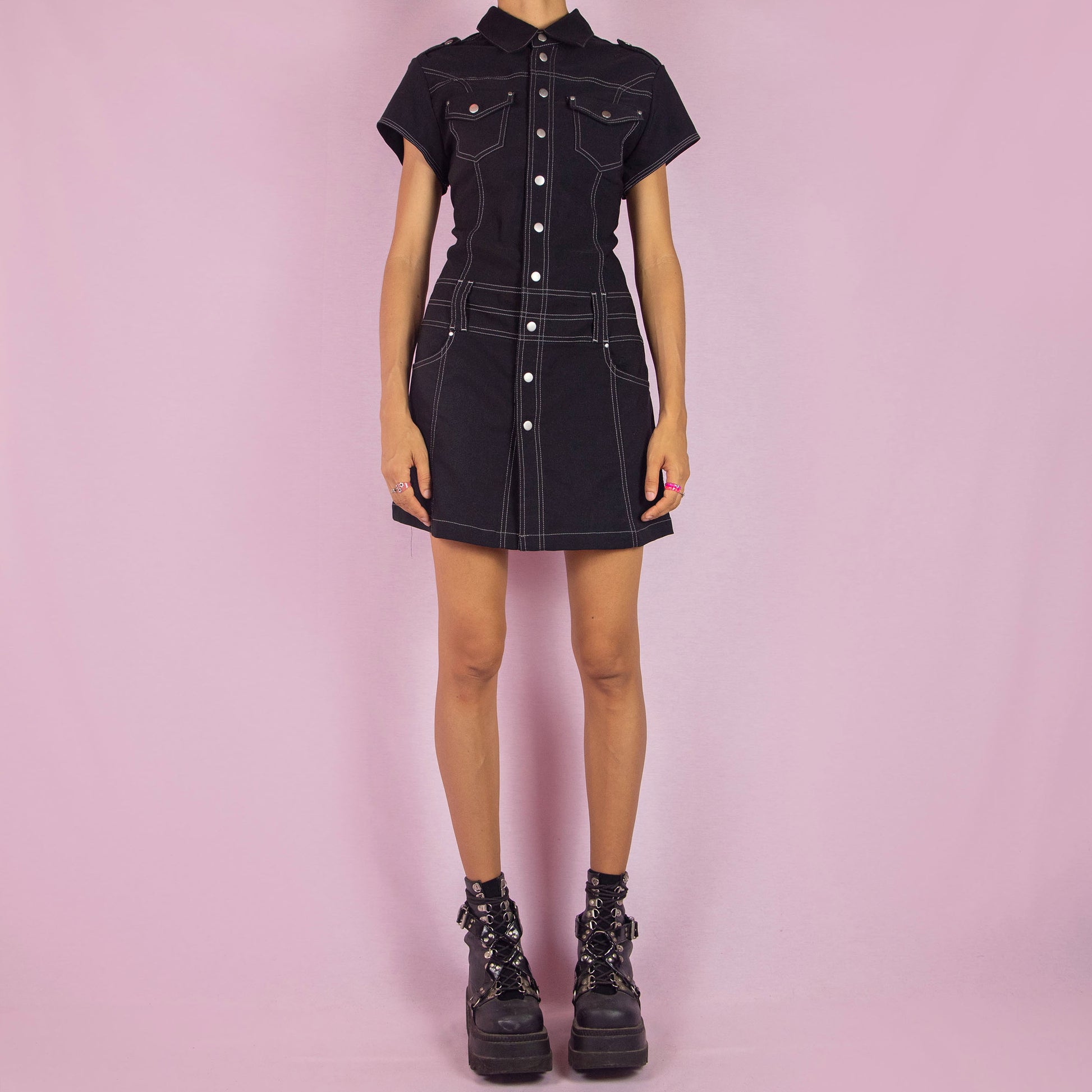 The Vintage 90s Black Utility Mini Dress is a sleek, structured piece featuring a collared neckline, short sleeves, and snap button closure along the front. The dress is designed with contrast stitching and stretchy fabric. Chest pockets and belt loops add a utilitarian touch, while the fitted silhouette and mini length make it perfect for casual outings or night events.