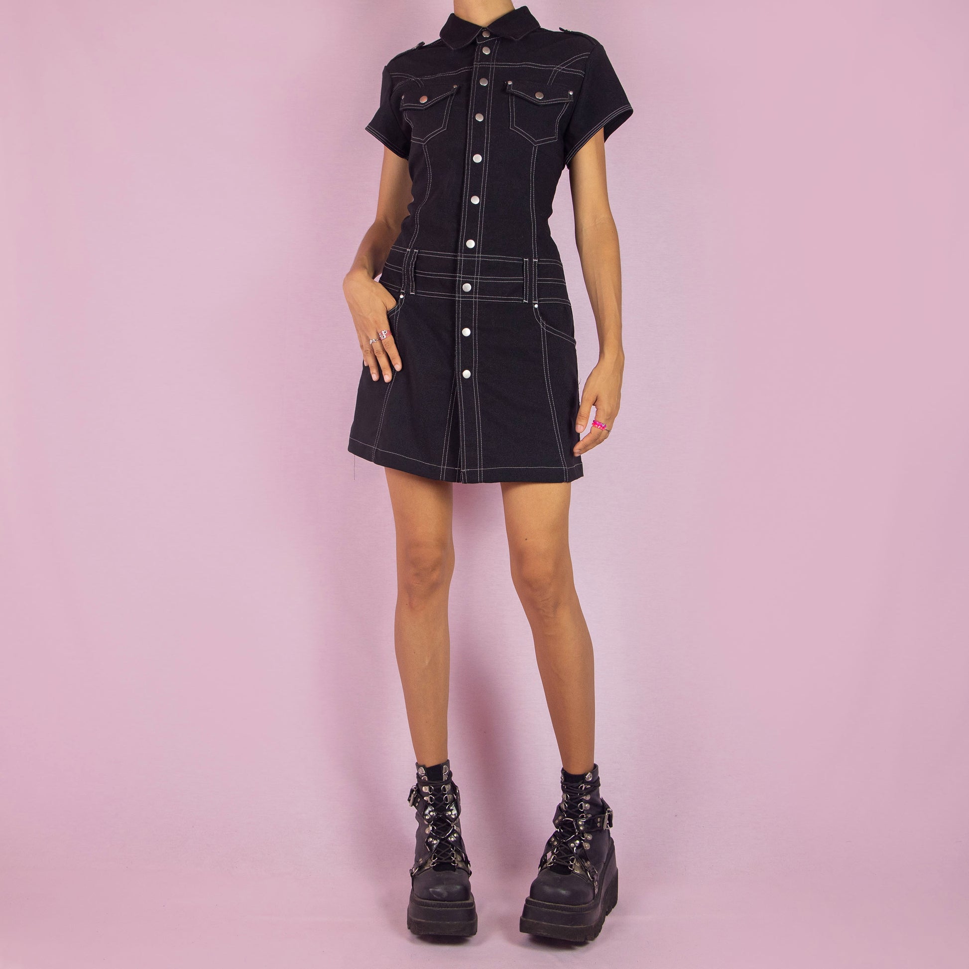 The Vintage 90s Black Utility Mini Dress is a sleek, structured piece featuring a collared neckline, short sleeves, and snap button closure along the front. The dress is designed with contrast stitching and stretchy fabric. Chest pockets and belt loops add a utilitarian touch, while the fitted silhouette and mini length make it perfect for casual outings or night events.