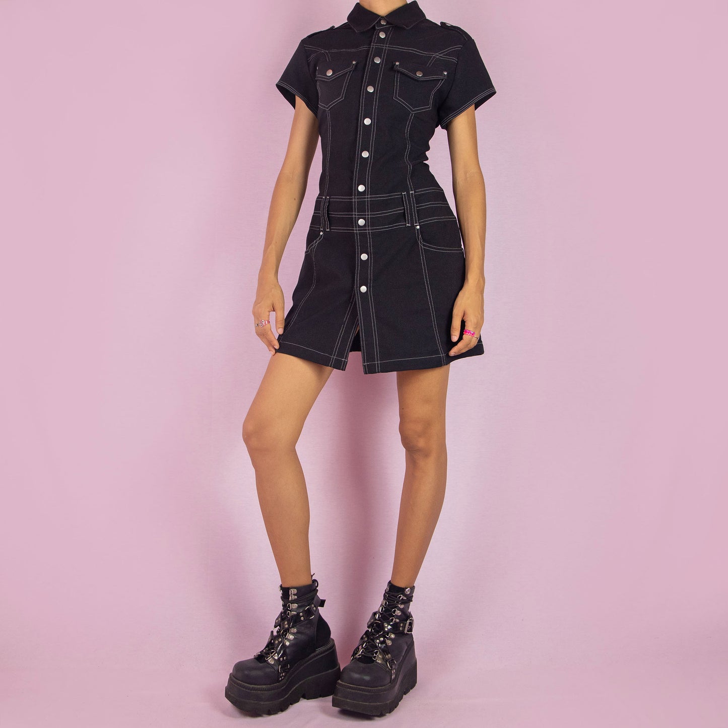 The Vintage 90s Black Utility Mini Dress is a sleek, structured piece featuring a collared neckline, short sleeves, and snap button closure along the front. The dress is designed with contrast stitching and stretchy fabric. Chest pockets and belt loops add a utilitarian touch, while the fitted silhouette and mini length make it perfect for casual outings or night events.