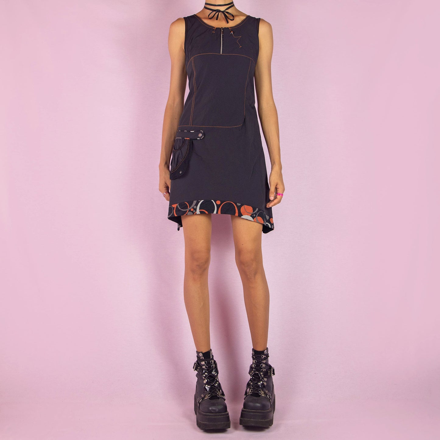 The Y2K Black Sleeveless Mini Dress is a vintage 2000s piece featuring a scoop neckline and a fitted silhouette made from stretchy fabric. This unique dress includes a zip-up front, a side pocket, and contrast stitching. It also features an asymmetric pointed hem with a geometric print trim. Perfect for parties and night events.