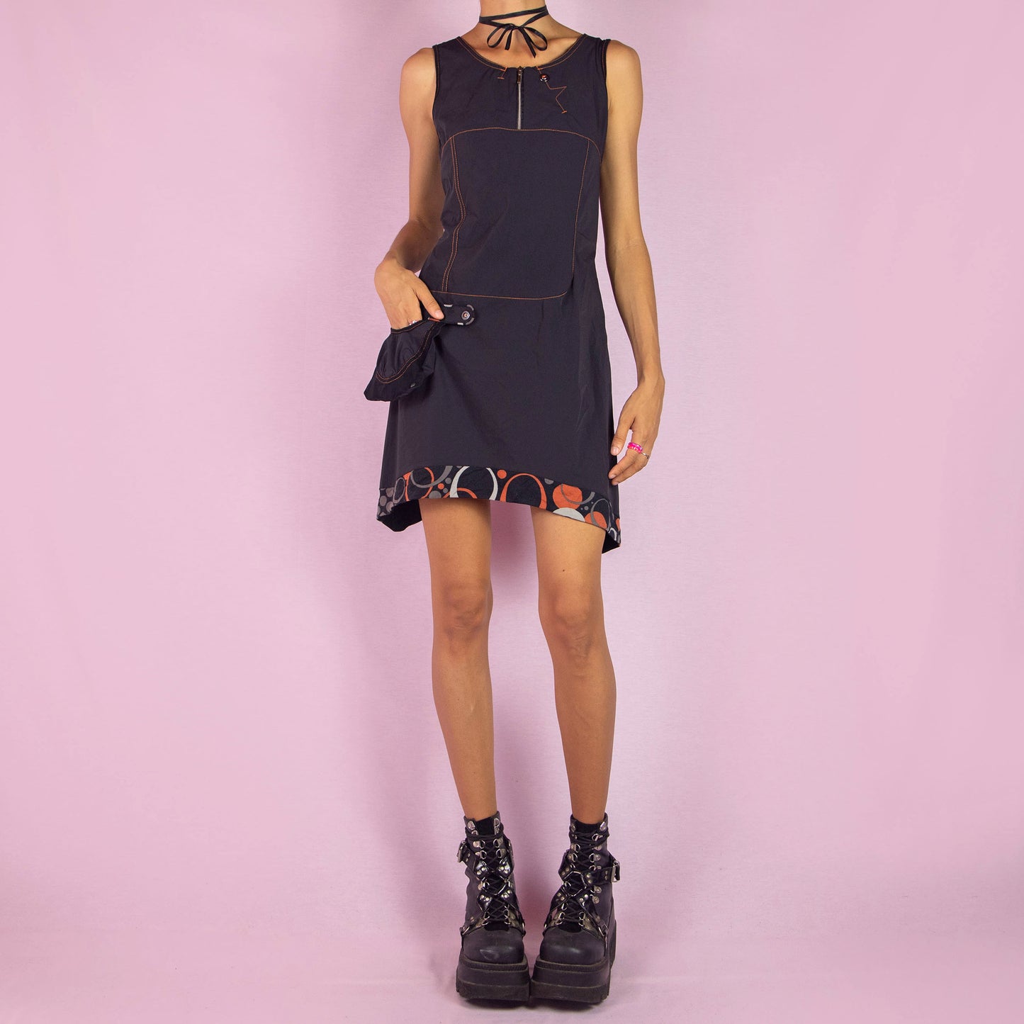 The Y2K Black Sleeveless Mini Dress is a vintage 2000s piece featuring a scoop neckline and a fitted silhouette made from stretchy fabric. This unique dress includes a zip-up front, a side pocket, and contrast stitching. It also features an asymmetric pointed hem with a geometric print trim. Perfect for parties and night events.