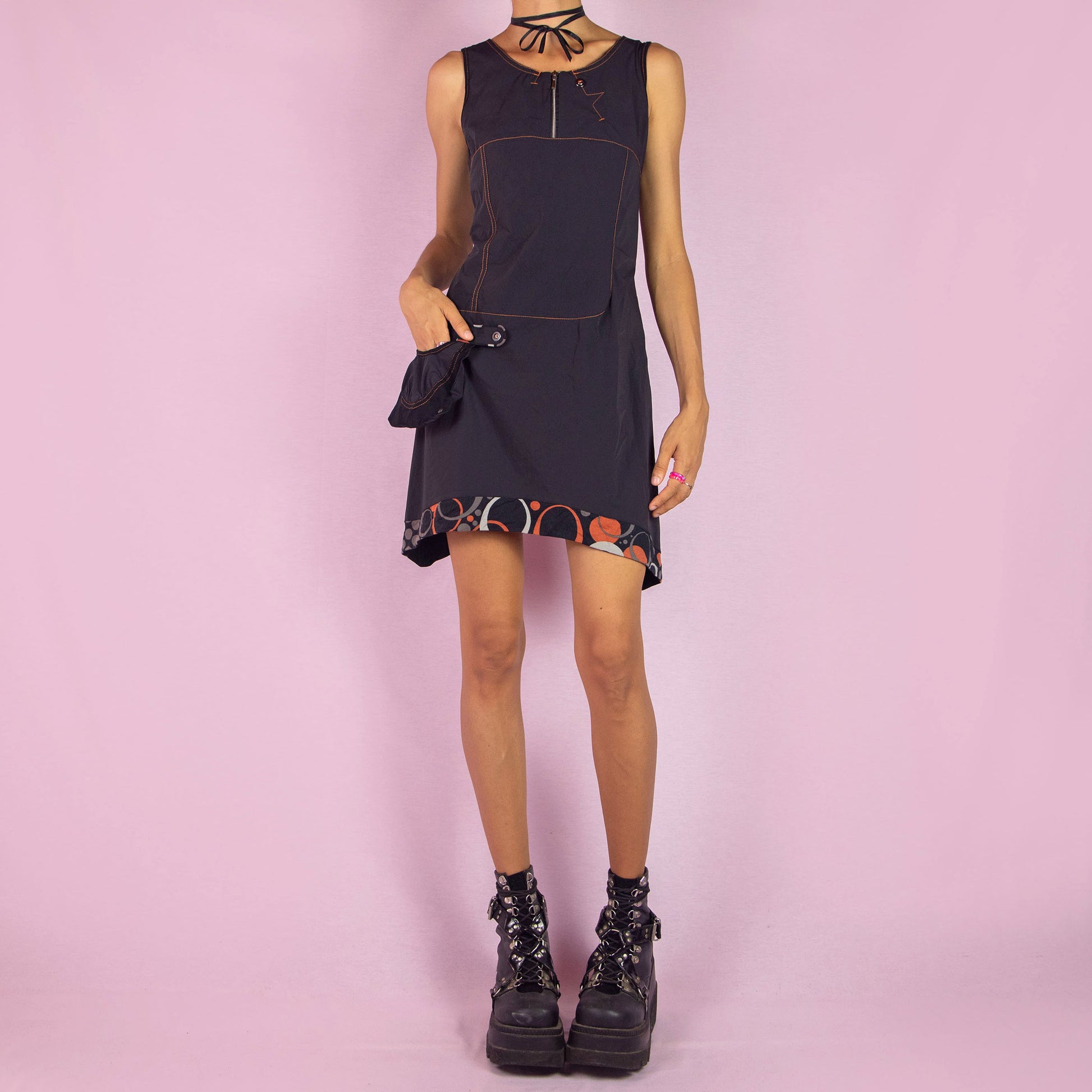 The Y2K Black Sleeveless Mini Dress is a vintage 2000s piece featuring a scoop neckline and a fitted silhouette made from stretchy fabric. This unique dress includes a zip-up front, a side pocket, and contrast stitching. It also features an asymmetric pointed hem with a geometric print trim. Perfect for parties and night events.