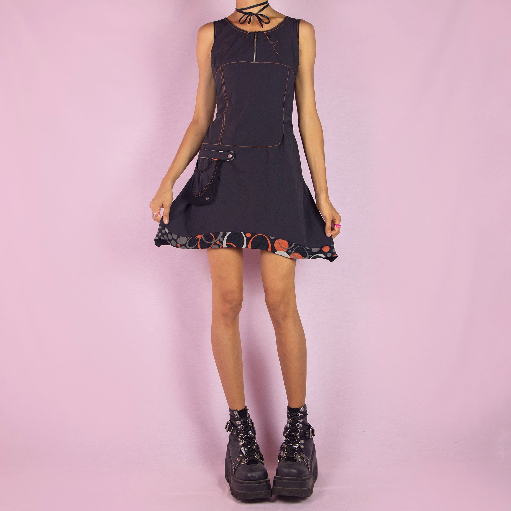 The Y2K Black Sleeveless Mini Dress is a vintage 2000s piece featuring a scoop neckline and a fitted silhouette made from stretchy fabric. This unique dress includes a zip-up front, a side pocket, and contrast stitching. It also features an asymmetric pointed hem with a geometric print trim. Perfect for parties and night events.