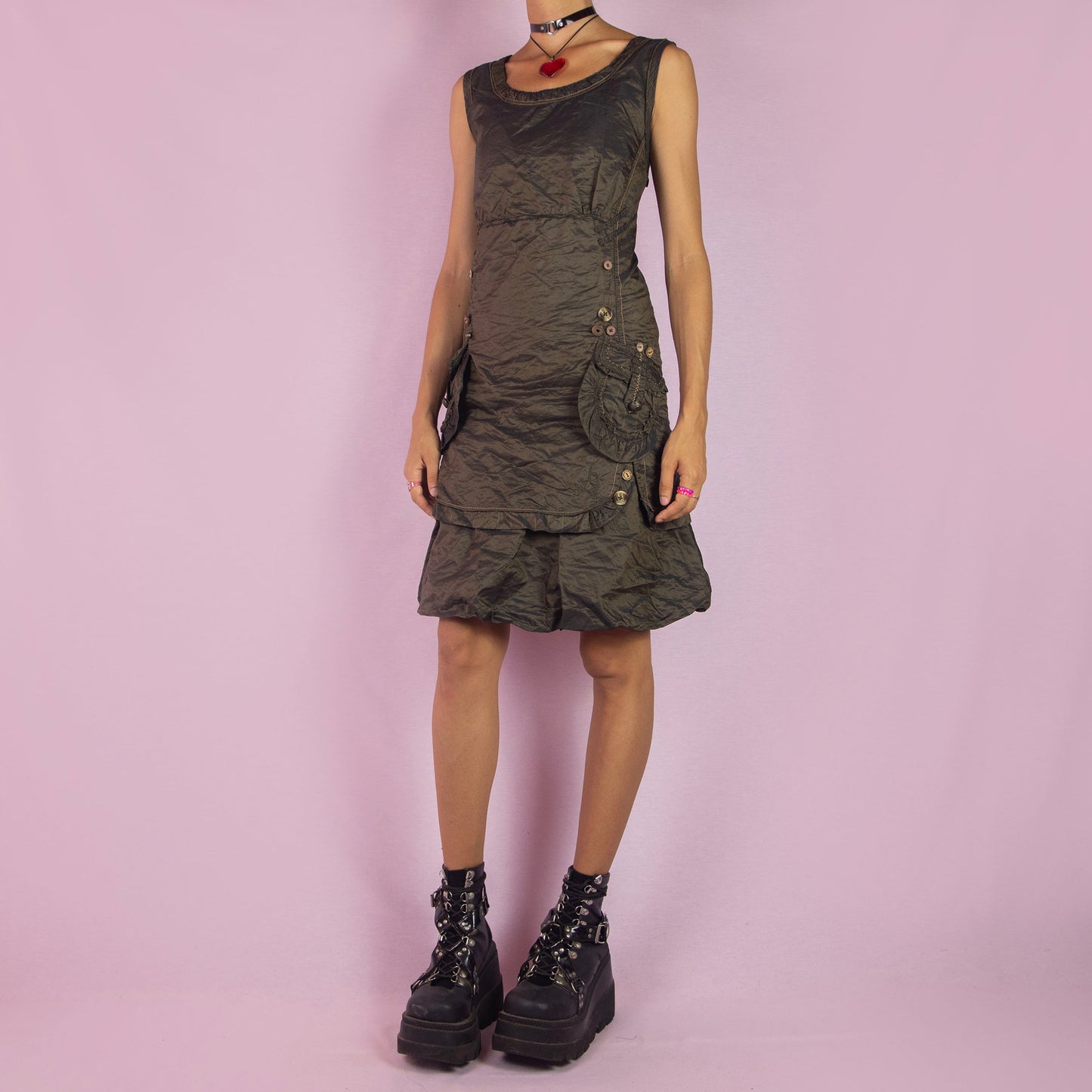 The Vintage 90s Layered Sleeveless Dress is a unique piece featuring a scoop neckline and an earthy olive-green tone. Designed with textured fabric, this dress includes a side zipper closure and detailed button accents along the sides and pockets. The layered skirt adds dimension, making it ideal for formal events or night parties.