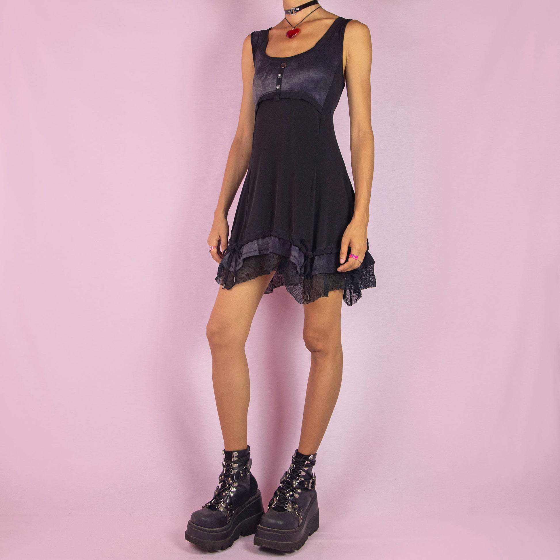 The Y2K Sleeveless Black Mini Dress is a vintage 2000s piece featuring a sleeveless design with a scoop neckline. Made from stretchy fabric, this dress has a buttoned bodice detail and a subtle tie-dye print across the top. The layered ruffle hem with mesh accents and adjustable drawstring ties adds a customizable fit. Perfect for summer casual outings or night parties.