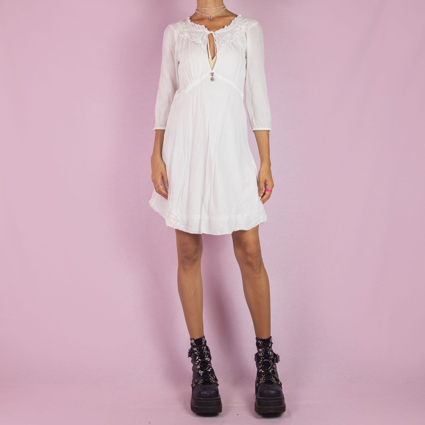 The Y2K White Tie Front Mini Dress is a vintage 2000s piece featuring a keyhole neckline and delicate embroidered floral designs along the bodice. Made from soft cotton, it has 3/4-length sleeves and a flowy A-line silhouette. Perfect for casual summer events or a romantic boho-inspired outfit.