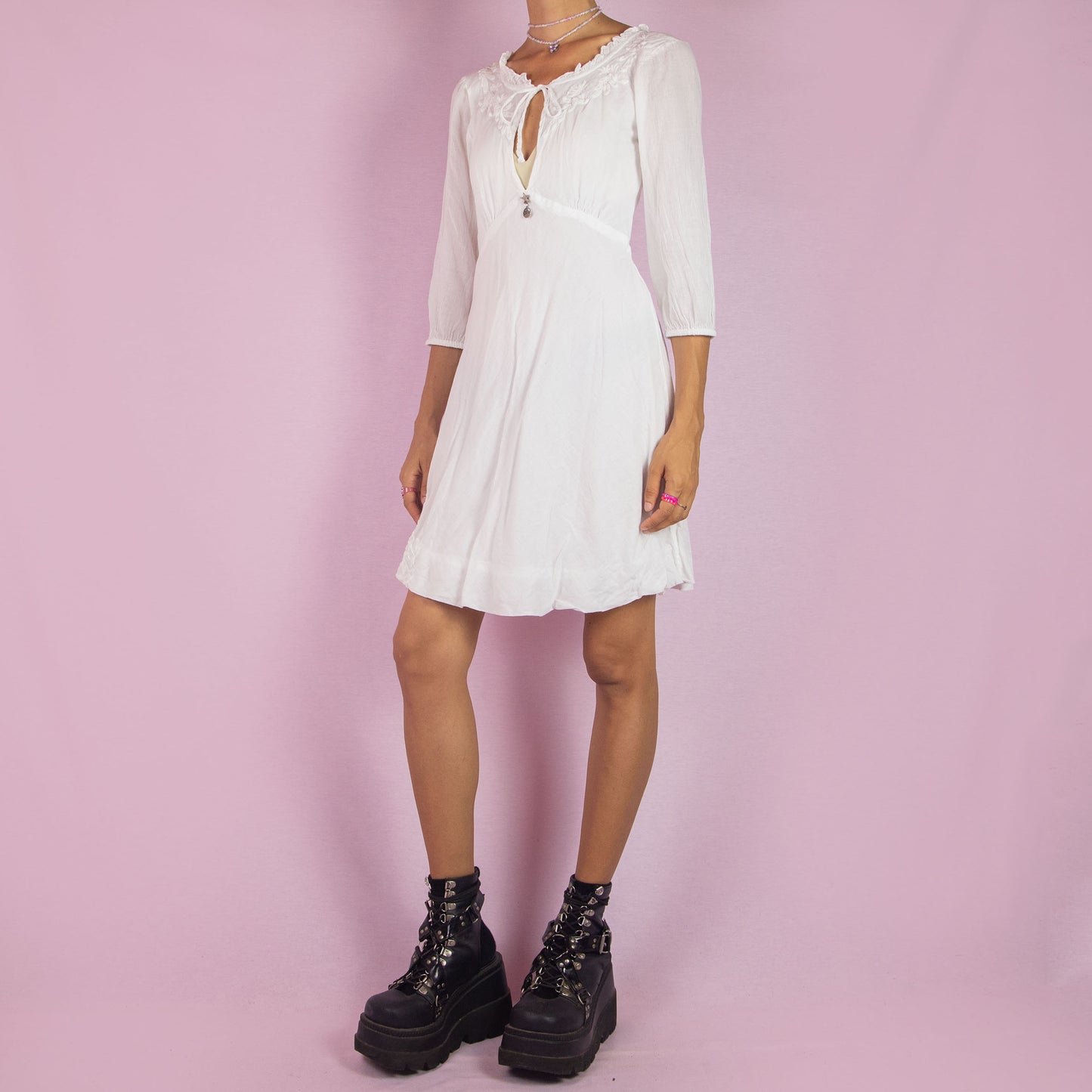 The Y2K White Tie Front Mini Dress is a vintage 2000s piece featuring a keyhole neckline and delicate embroidered floral designs along the bodice. Made from soft cotton, it has 3/4-length sleeves and a flowy A-line silhouette. Perfect for casual summer events or a romantic boho-inspired outfit.