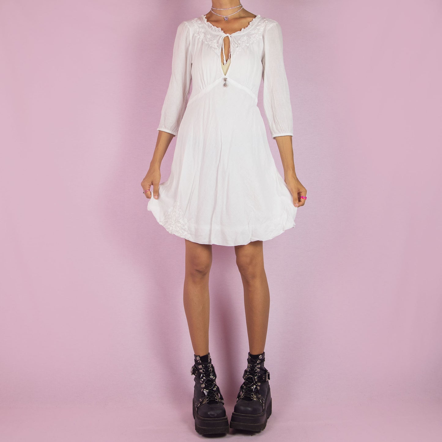 The Y2K White Tie Front Mini Dress is a vintage 2000s piece featuring a keyhole neckline and delicate embroidered floral designs along the bodice. Made from soft cotton, it has 3/4-length sleeves and a flowy A-line silhouette. Perfect for casual summer events or a romantic boho-inspired outfit.