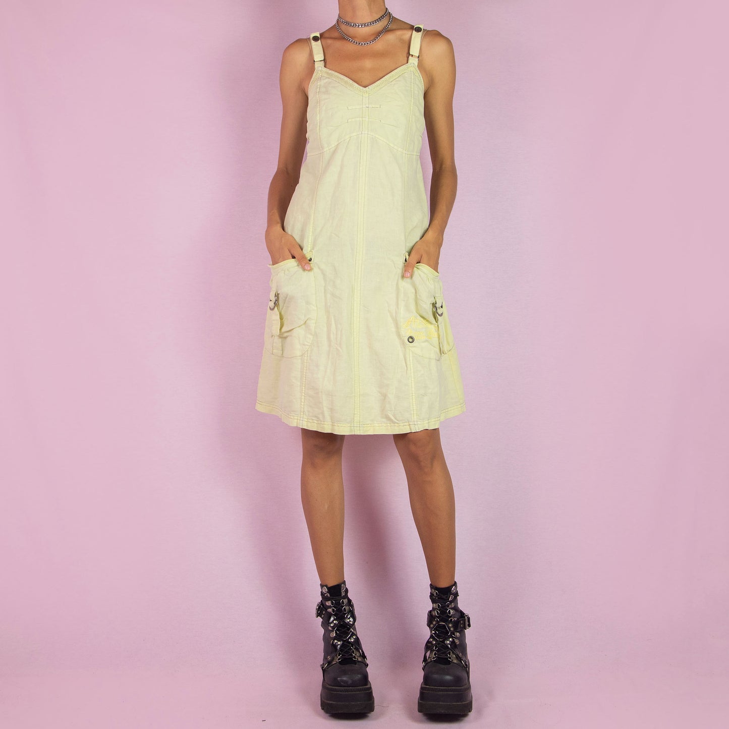 The Y2K Cotton Linen Midi Dress is a vintage 2000s piece featuring a sweetheart neckline, adjustable straps, and a back zipper closure. Made from a breathable pastel light yellow cotton-linen blend, this dress has utility-inspired details with side pockets, buckle accents, and a relaxed fit. Perfect for casual summer outings.