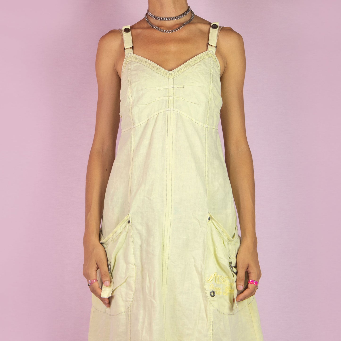 The Y2K Cotton Linen Midi Dress is a vintage 2000s piece featuring a sweetheart neckline, adjustable straps, and a back zipper closure. Made from a breathable pastel light yellow cotton-linen blend, this dress has utility-inspired details with side pockets, buckle accents, and a relaxed fit. Perfect for casual summer outings.