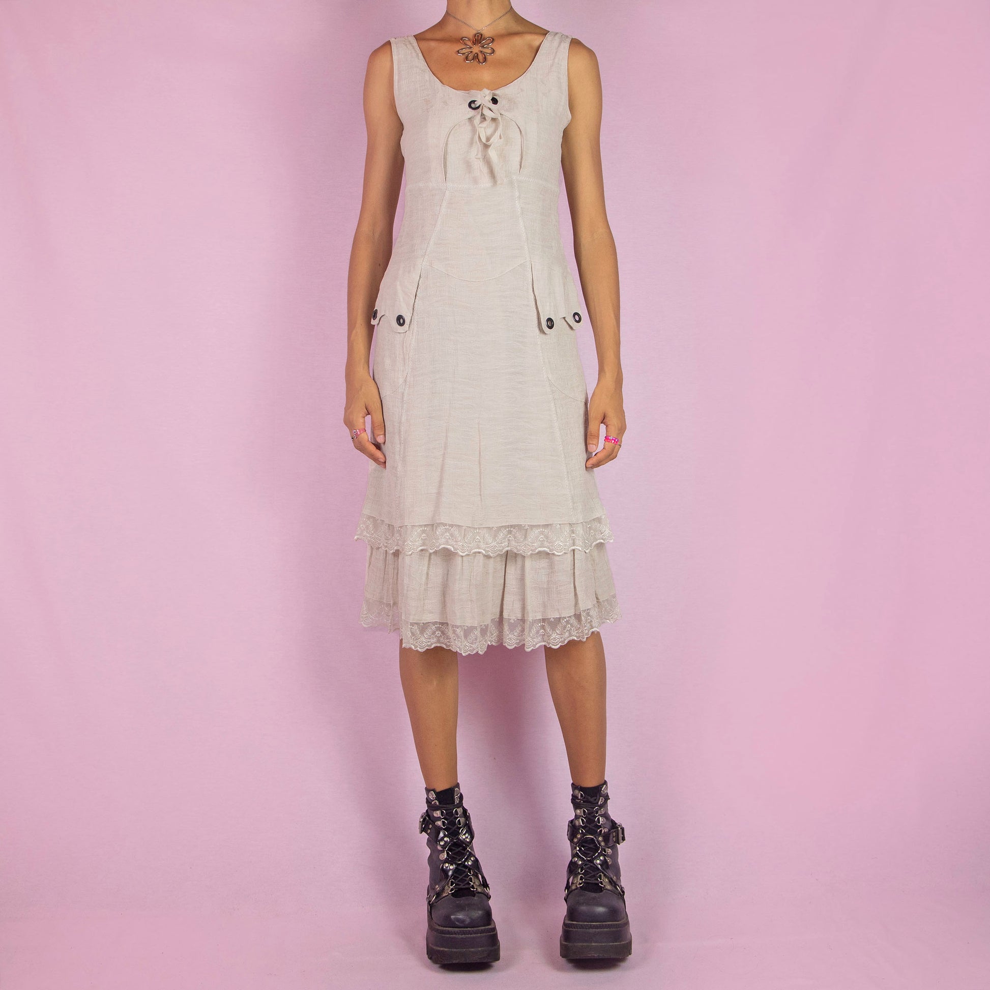 The Y2K Beige Sleeveless Midi Dress is a vintage 2000s piece featuring a lace-up front detail and a side zipper closure. Made from a textured cotton blend, this dress offers a flattering silhouette with pockets, and a charming tiered lace hem. Perfect for casual summer outings or an elegant boho-inspired look.