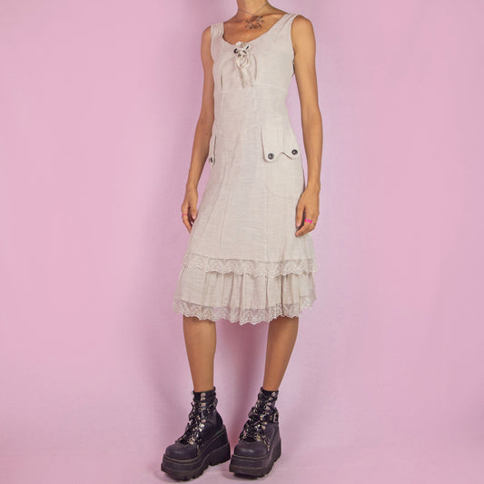 The Y2K Beige Sleeveless Midi Dress is a vintage 2000s piece featuring a lace-up front detail and a side zipper closure. Made from a textured cotton blend, this dress offers a flattering silhouette with pockets, and a charming tiered lace hem. Perfect for casual summer outings or an elegant boho-inspired look.