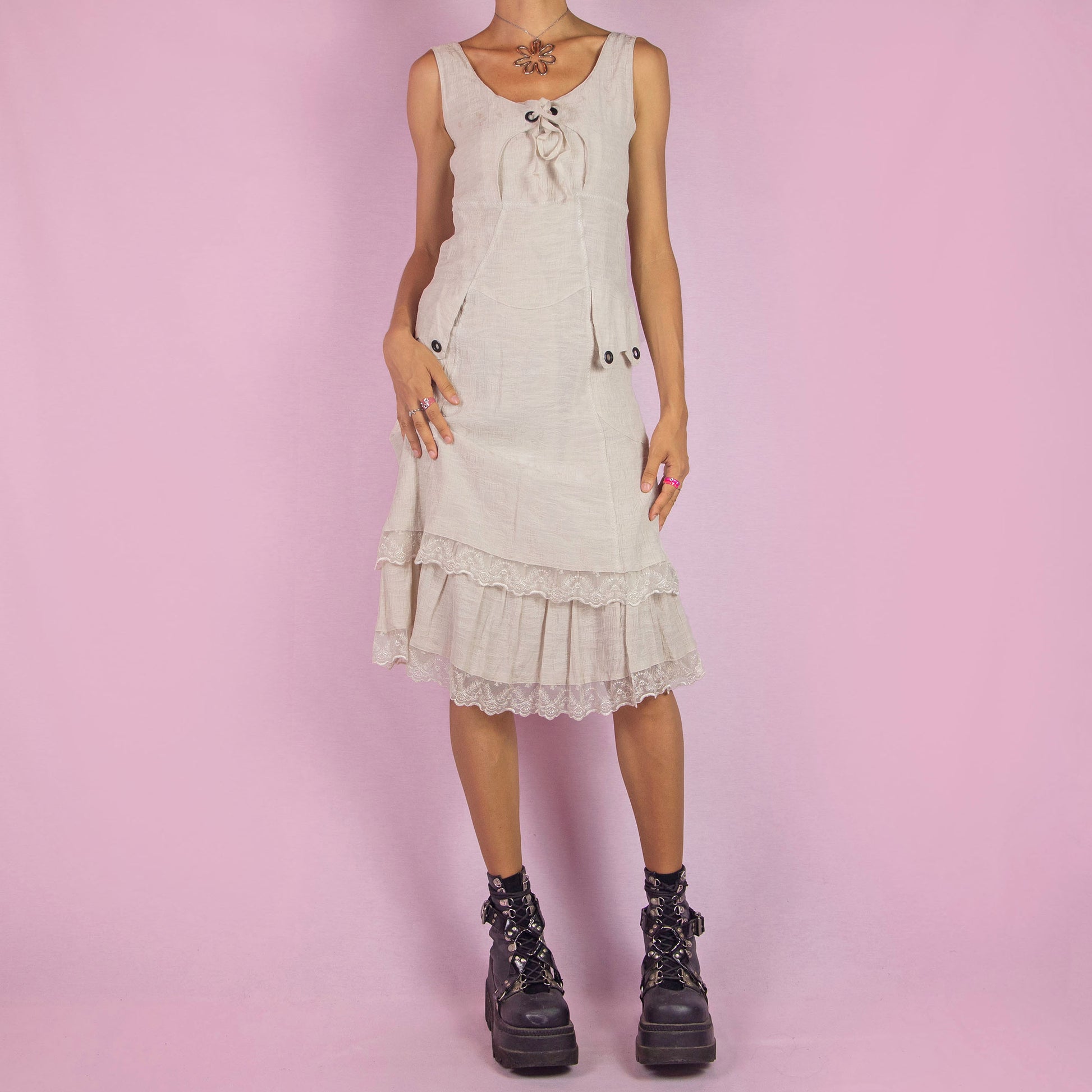 The Y2K Beige Sleeveless Midi Dress is a vintage 2000s piece featuring a lace-up front detail and a side zipper closure. Made from a textured cotton blend, this dress offers a flattering silhouette with pockets, and a charming tiered lace hem. Perfect for casual summer outings or an elegant boho-inspired look.