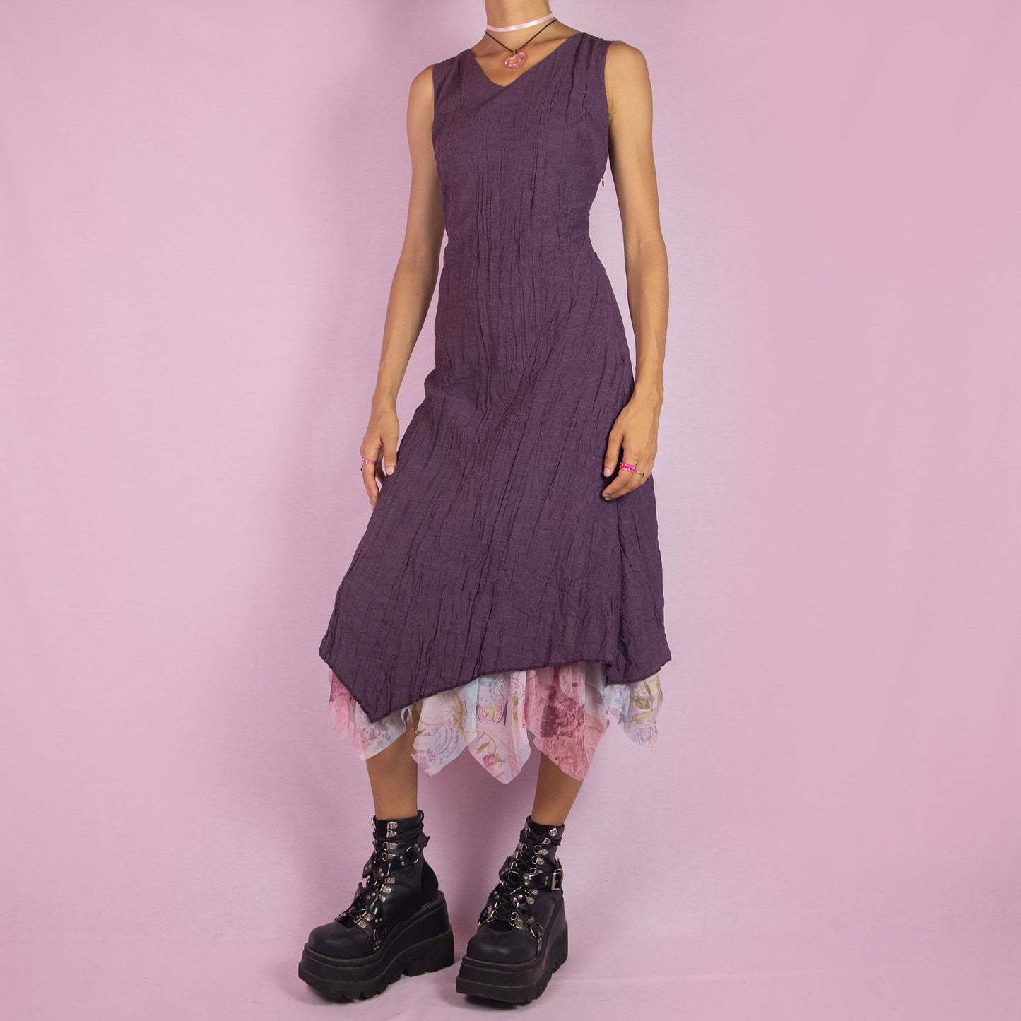 The Y2K Purple Sleeveless Midi Dress is a vintage 2000s piece featuring a crinkled, textured fabric, a V-neckline, and a side zipper closure. The dress also has a layered asymmetric hemline with an abstract floral print mesh sheer underlayer peeking out. Perfect for summer outings and elegant parties.