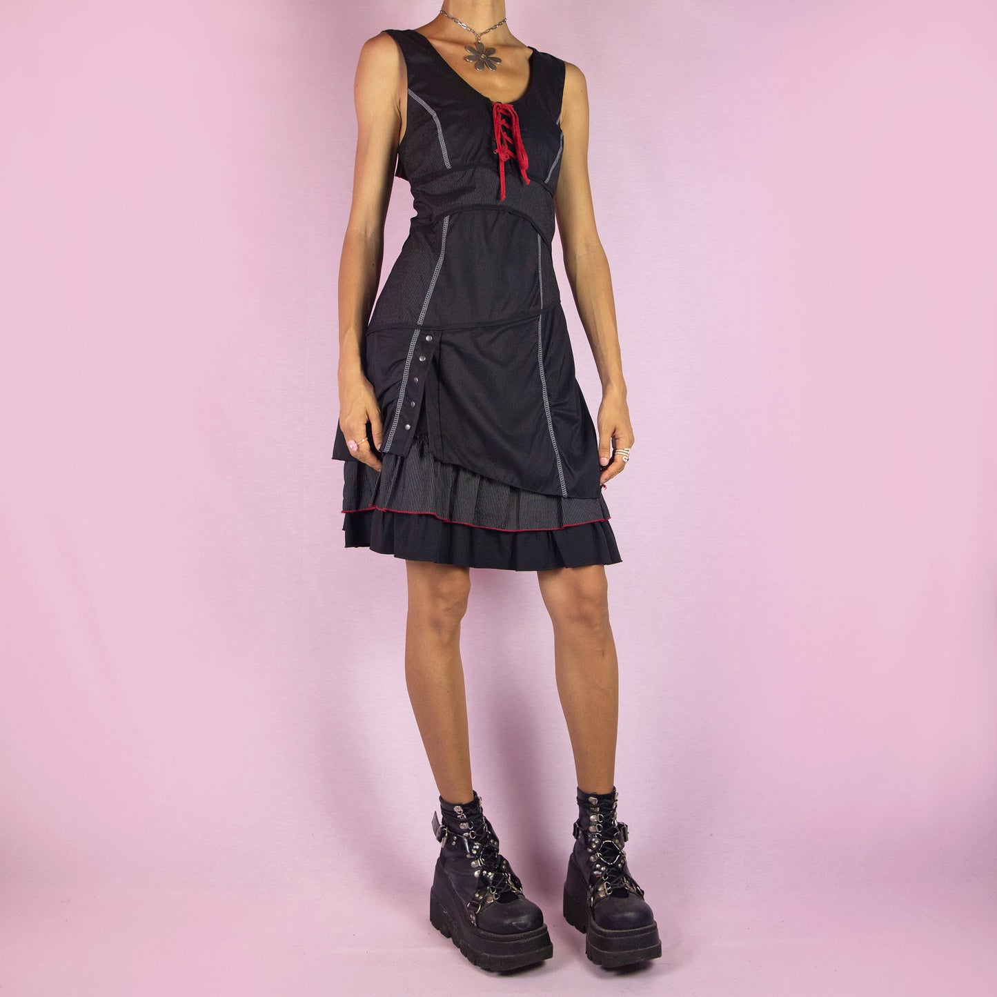 The Y2K Black Layered Lace-Up Dress is a vintage 2000s mini dress featuring a sleeveless design, scoop neckline, and stretchy fabric. It showcases a red lace-up detail at the bodice, contrasting stitching throughout, and a side zipper closure. The layered skirt, with its pinstripe and solid panels, button accents, and ruffle hem, adds a touch of whimsy. Perfect for night events and parties.