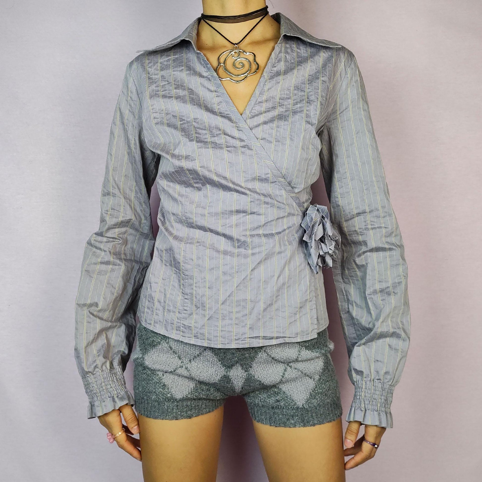 The Y2K Gray Tie Wrap Blouse is a vintage 2000s top featuring a chic wrap-around design with a side tie and delicate pinstripe pattern. Its tailored fit is complemented by a collared neckline and puffed sleeves with gathered cuffs.