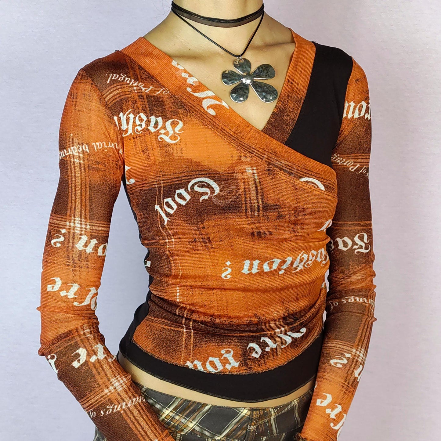 The Y2K Print Mesh Wrap Top is a vintage 2000s piece featuring a vibrant orange and brown design with bold typography prints. Crafted from a lightweight mesh fabric, it has a wrap-style front that accentuates the silhouette and long sleeves