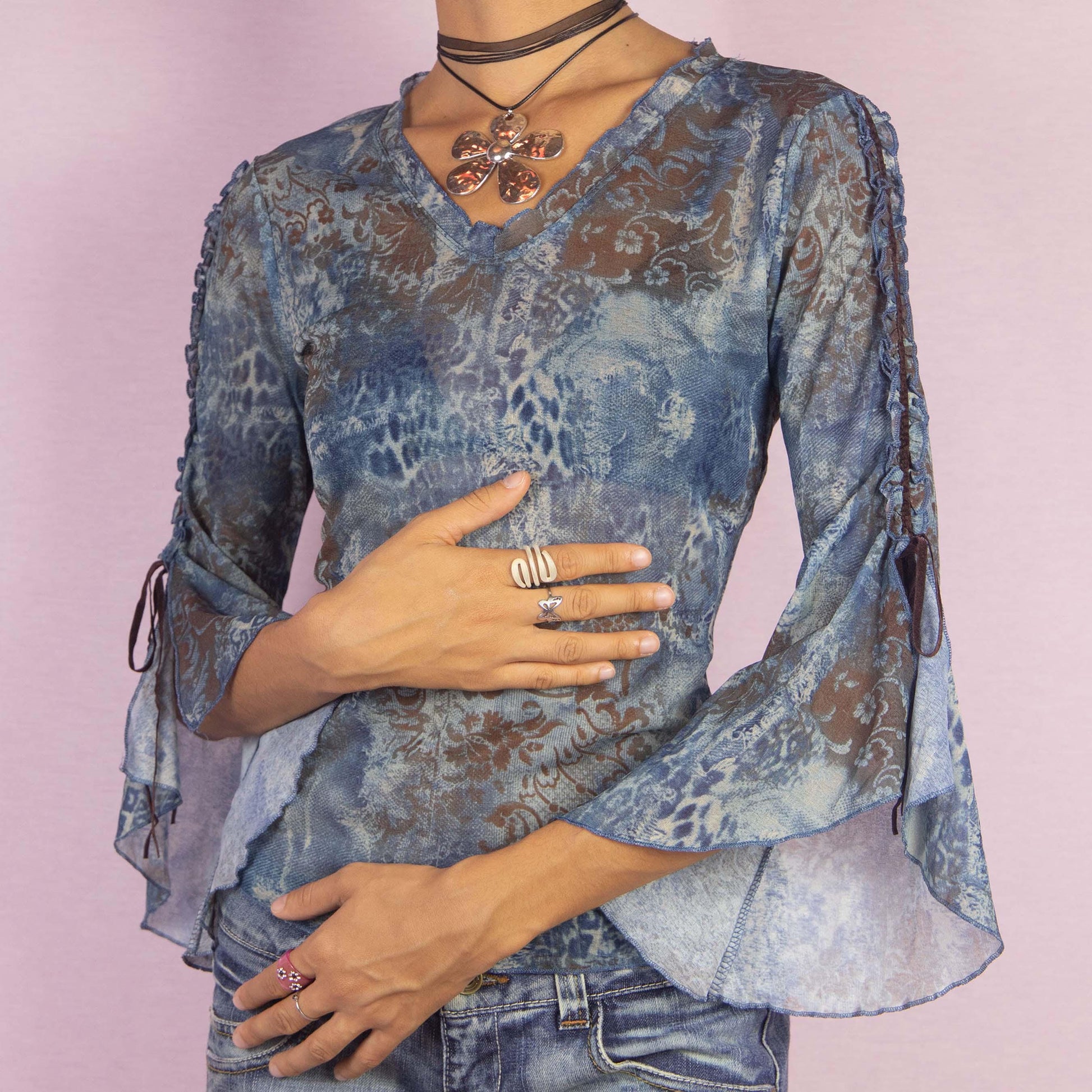 The Y2K Bell Sleeve Mesh Top is a vintage 2000s blouse featuring a semi-sheer fabric with an abstract, blue-toned pattern and intricate baroque-inspired prints. This unique piece boasts dramatic flared sleeves with tie-up details