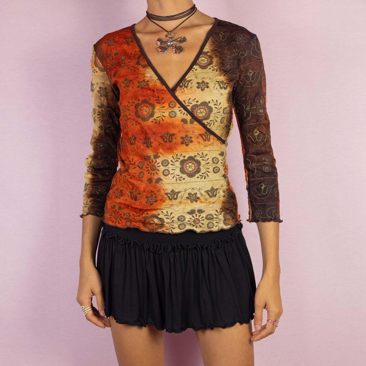 The Y2K Tie Dye Wrap Top is a vintage 2000s shirt featuring a tie-dye-inspired gradient in earthy orange and brown hues, complemented by mandala-inspired patterns. This top showcases a flattering wrap-style silhouette with a deep V-neckline