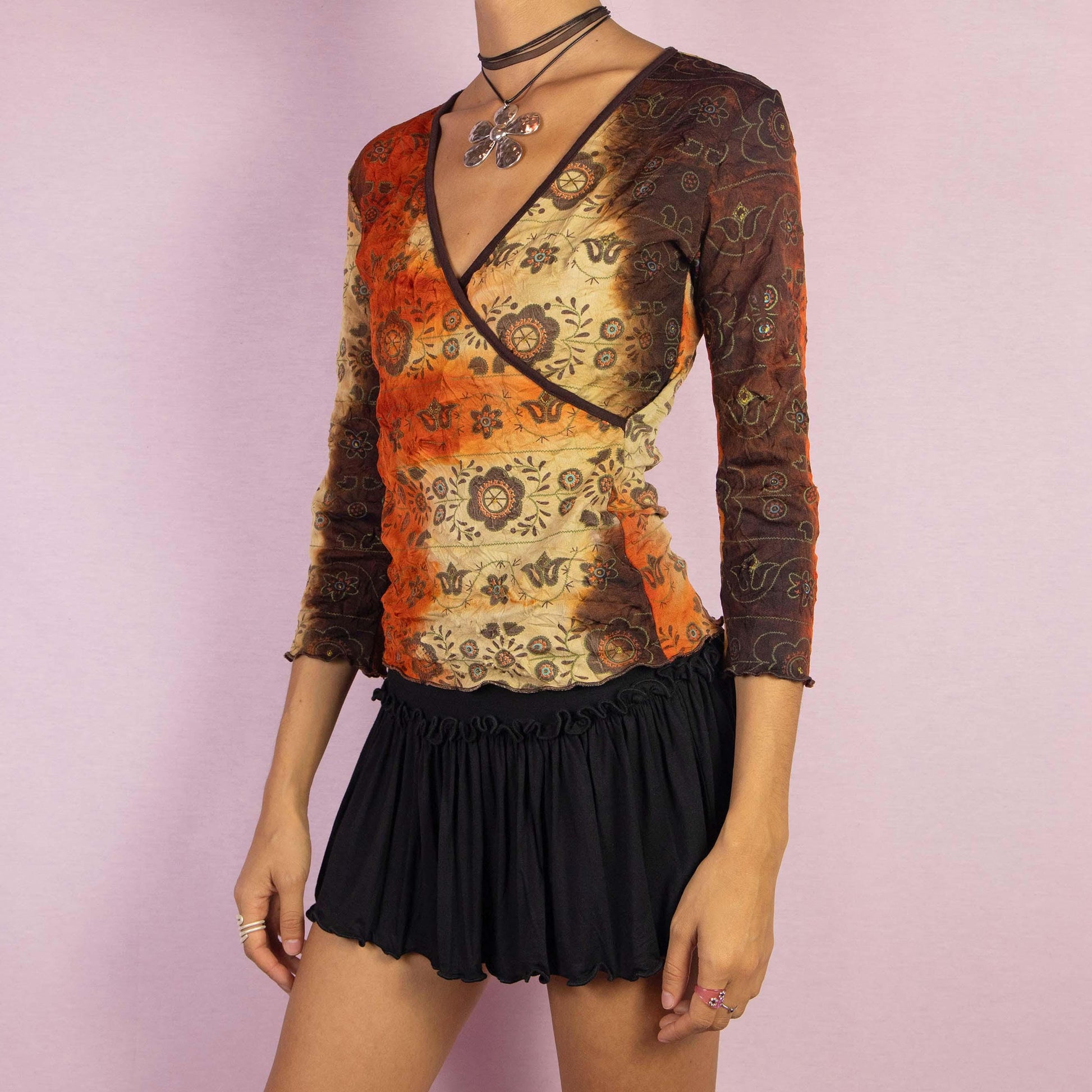 The Y2K Tie Dye Wrap Top is a vintage 2000s shirt featuring a tie-dye-inspired gradient in earthy orange and brown hues, complemented by mandala-inspired patterns. This top showcases a flattering wrap-style silhouette with a deep V-neckline