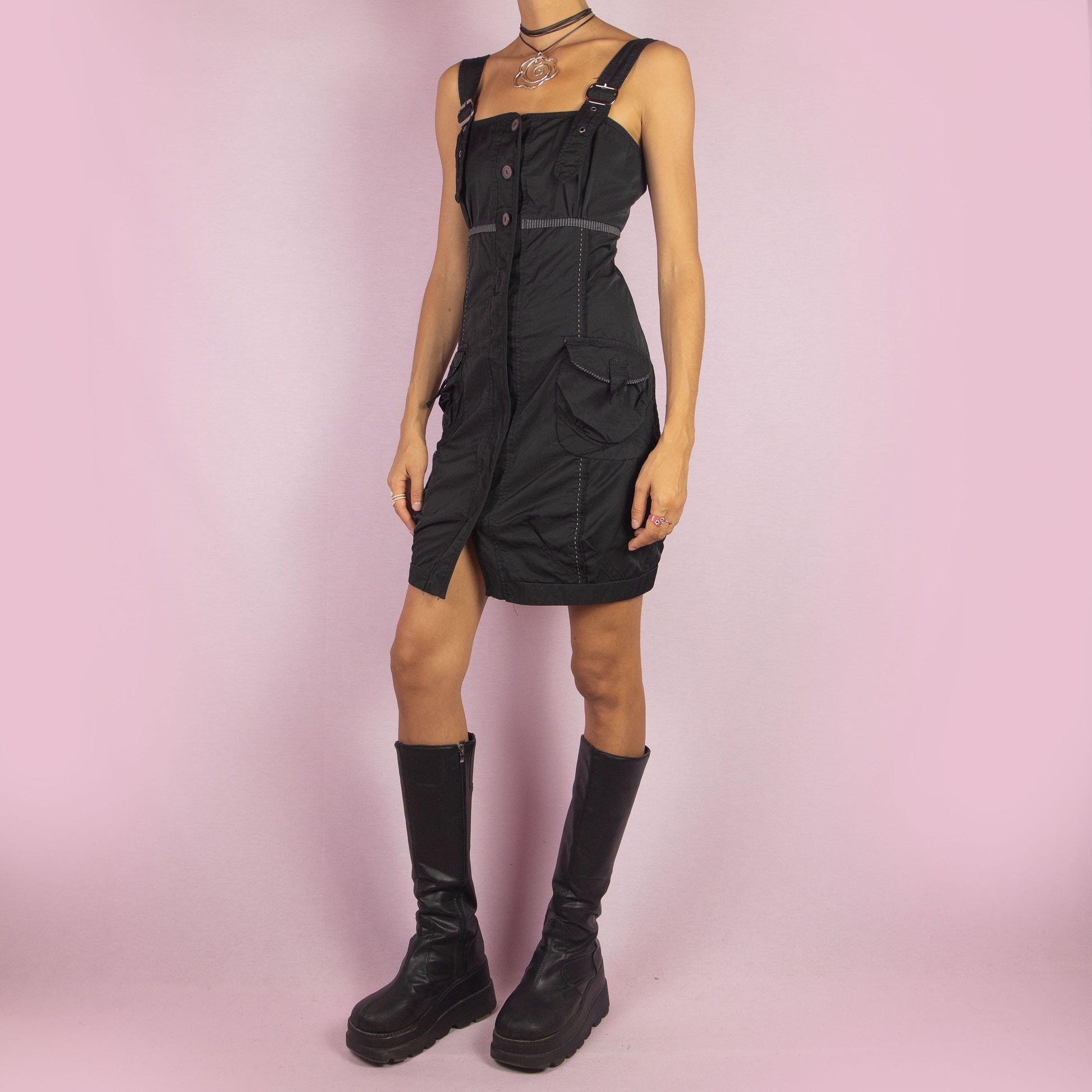 The Y2K Buckle Strap Black Mini Dress is a vintage 2000s piece featuring adjustable buckle straps, a button-down front, and utility-style pockets. Designed with contrast stitching and a structured silhouette, this mini dress is perfect for daytime