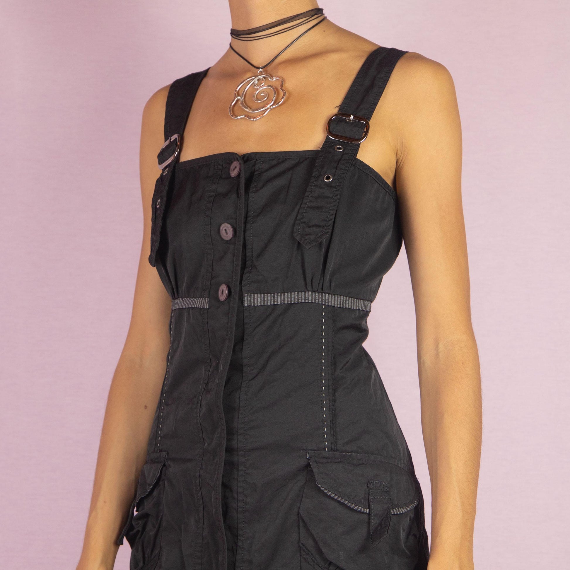 The Y2K Buckle Strap Black Mini Dress is a vintage 2000s piece featuring adjustable buckle straps, a button-down front, and utility-style pockets. Designed with contrast stitching and a structured silhouette, this mini dress is perfect for daytime