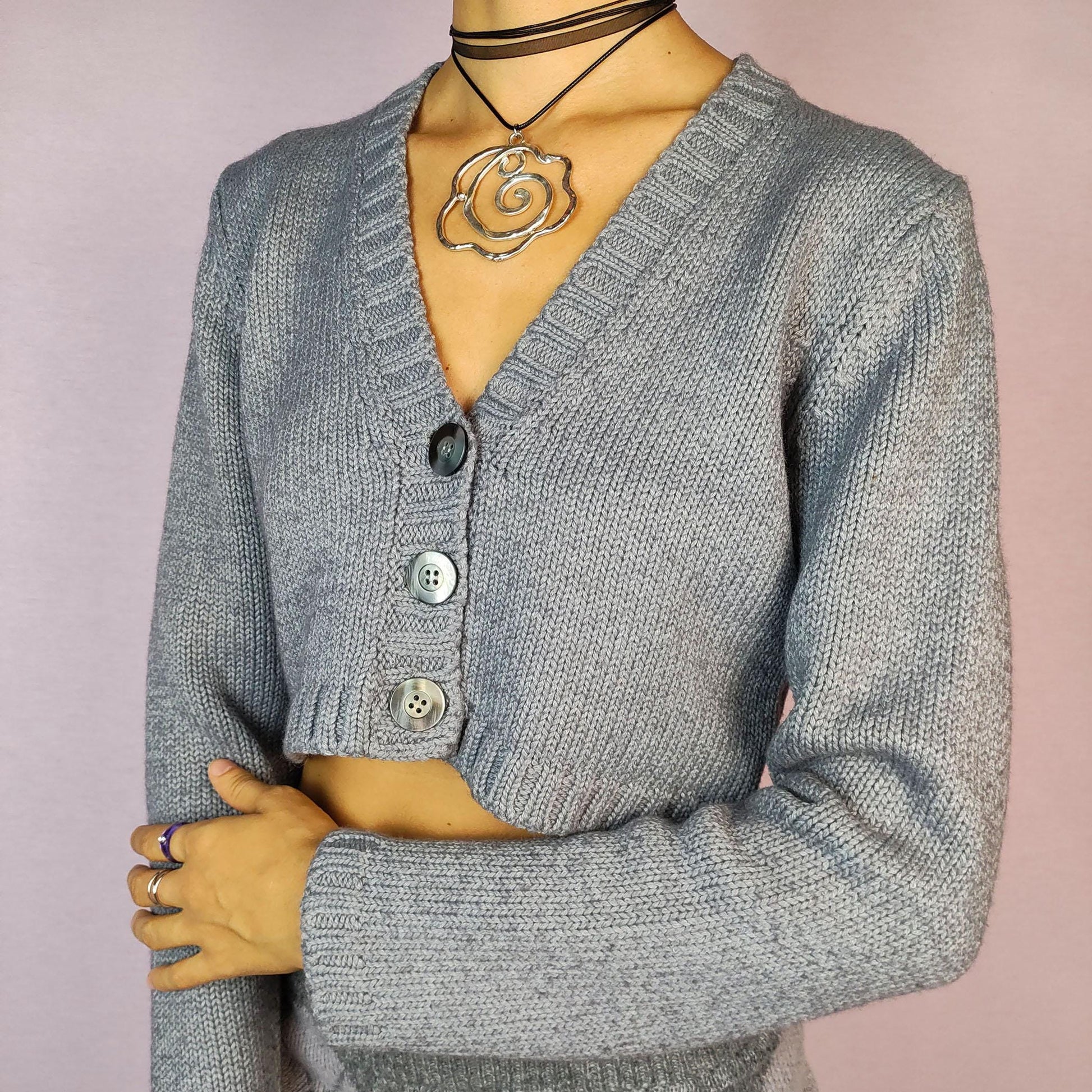 The Y2K Gray Cropped Cardigan is a vintage 2000s sweater featuring a classic V-neck design with a button-front closure, long sleeves, and ribbed detailing on the hem and cuffs. Crafted from a merino wool and acrylic blend