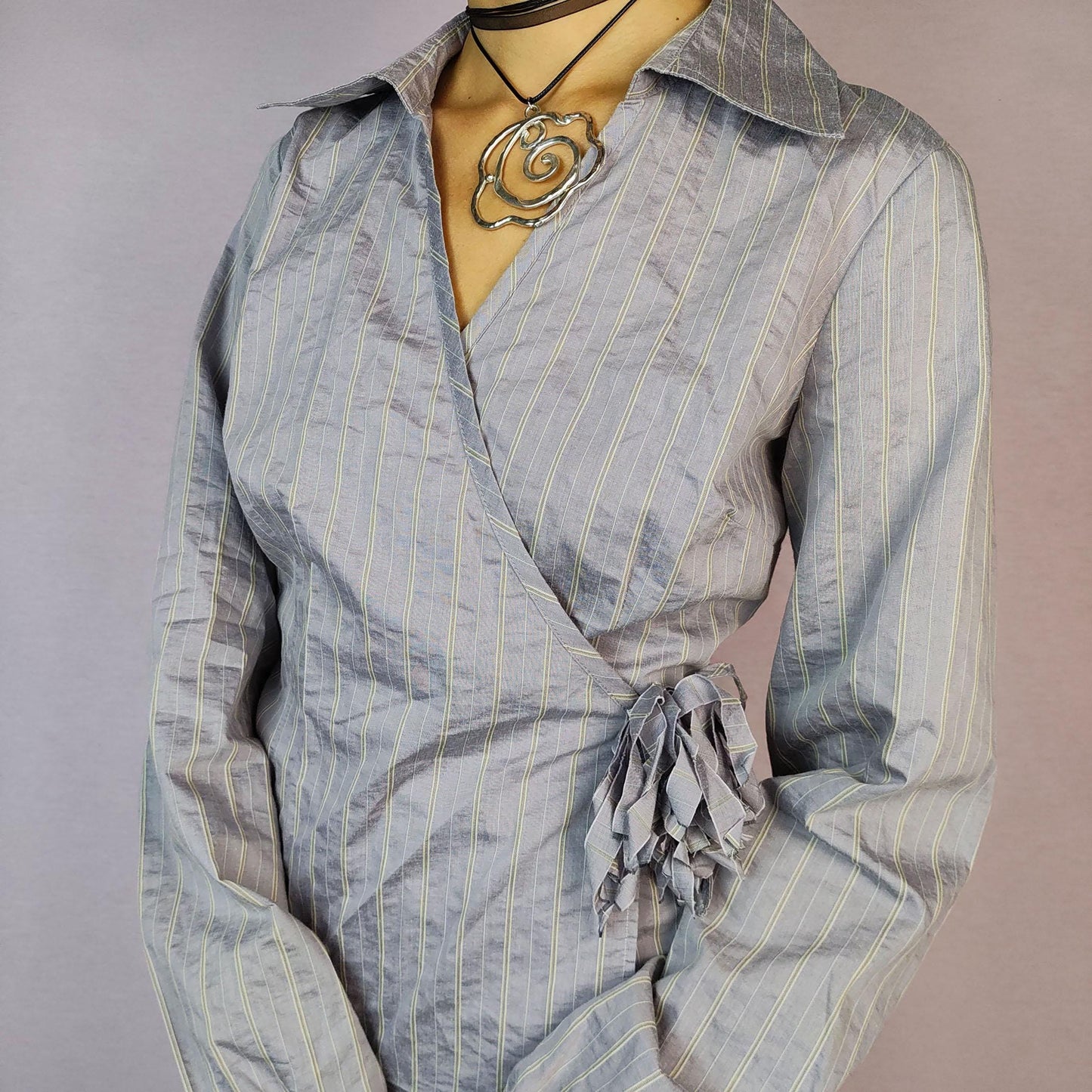 The Y2K Gray Tie Wrap Blouse is a vintage 2000s top featuring a chic wrap-around design with a side tie and delicate pinstripe pattern. Its tailored fit is complemented by a collared neckline and puffed sleeves with gathered cuffs.