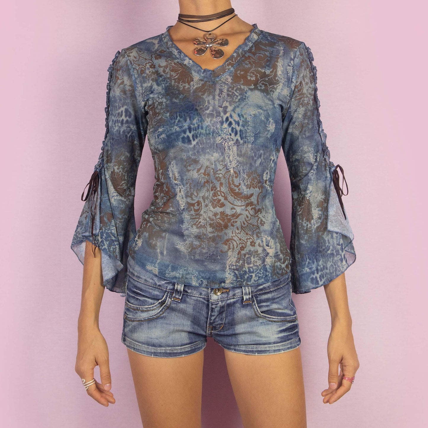 The Y2K Bell Sleeve Mesh Top is a vintage 2000s blouse featuring a semi-sheer fabric with an abstract, blue-toned pattern and intricate baroque-inspired prints. This unique piece boasts dramatic flared sleeves with tie-up details