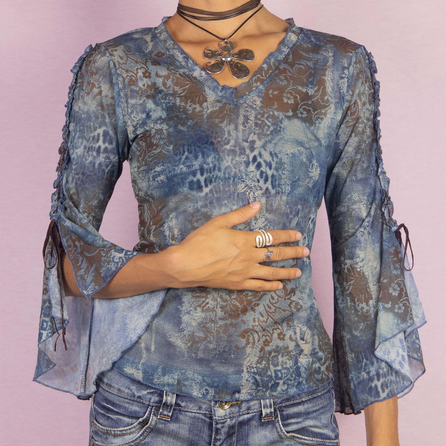 The Y2K Bell Sleeve Mesh Top is a vintage 2000s blouse featuring a semi-sheer fabric with an abstract, blue-toned pattern and intricate baroque-inspired prints. This unique piece boasts dramatic flared sleeves with tie-up details