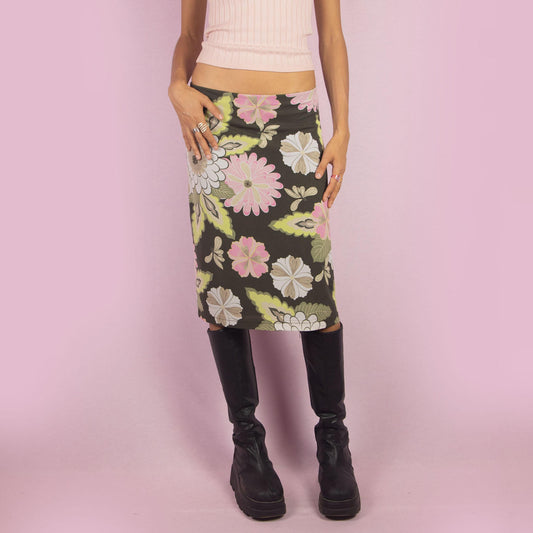 The Y2K Green Floral Mesh Skirt is a vintage 2000s piece featuring a floral print in soft pink, white, and khaki green tones on a semi-sheer mesh fabric. Its midi-length cut and elastic waist offer a flattering and comfortable fit