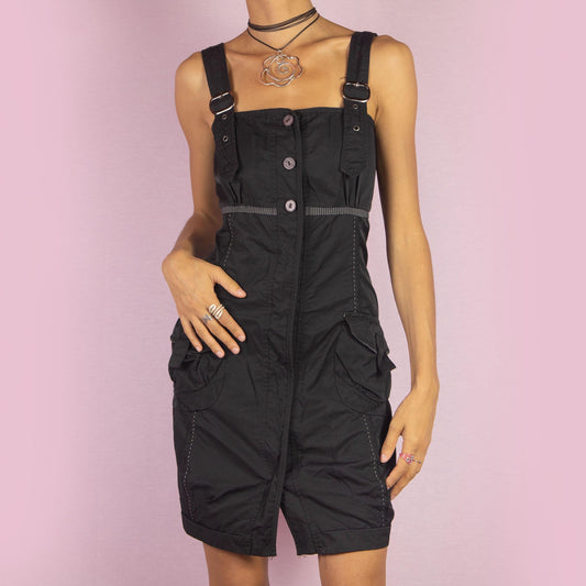 The Y2K Buckle Strap Black Mini Dress is a vintage 2000s piece featuring adjustable buckle straps, a button-down front, and utility-style pockets. Designed with contrast stitching and a structured silhouette, this mini dress is perfect for daytime